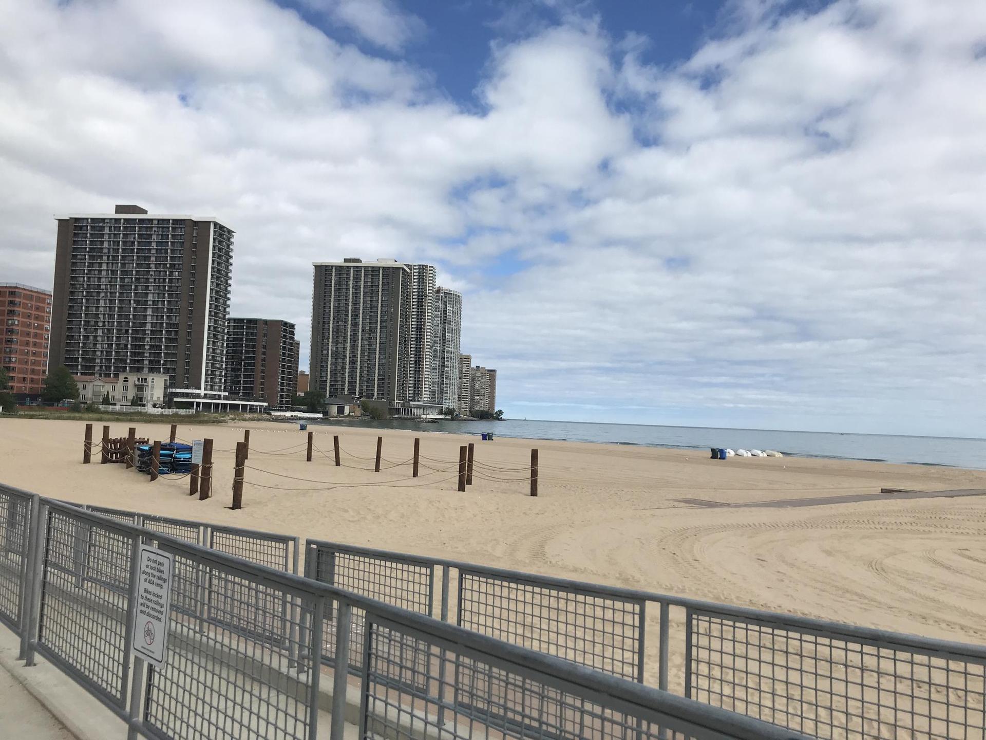 Empty sandy beach with tall buildings and calm water under cloudy sky.