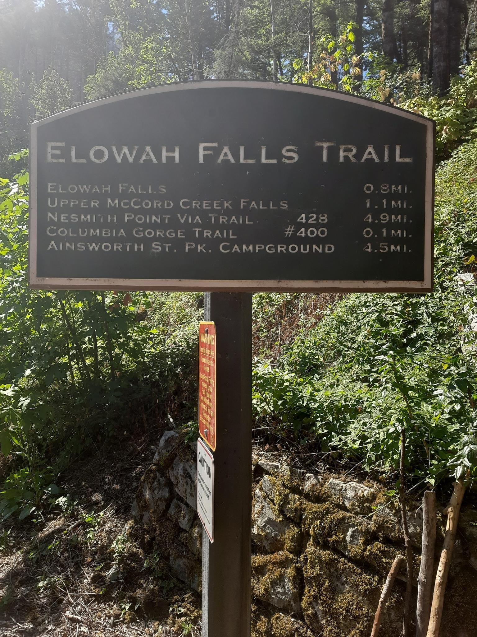 A dark trail sign with distances, surrounded by green foliage in sunlight. - Elowah and Upper McCord Creek Falls Trail