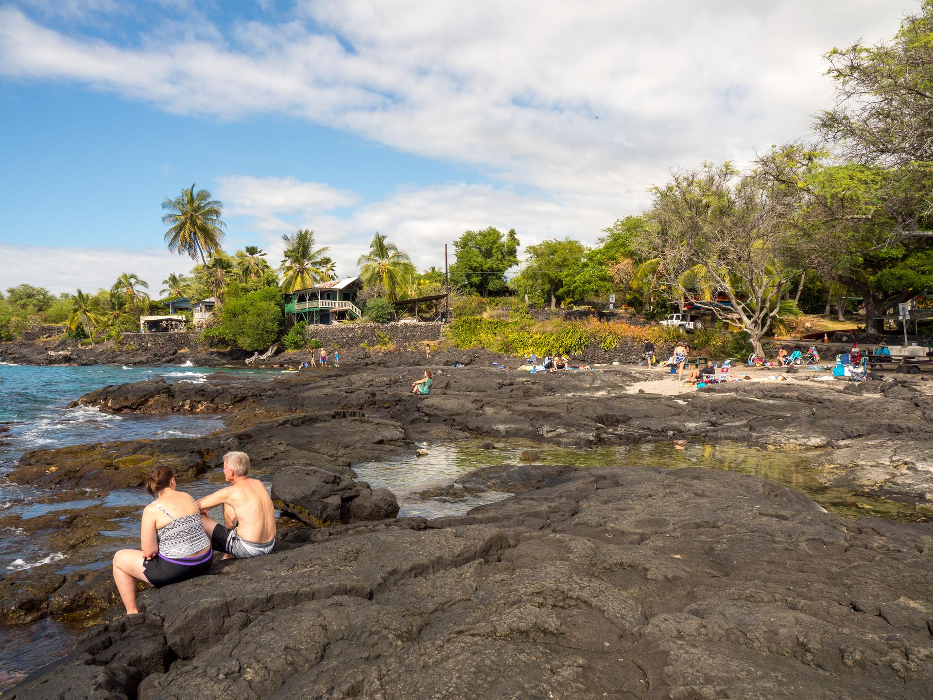 People relax on dark volcanic rocks by the ocean under a sunny sky.