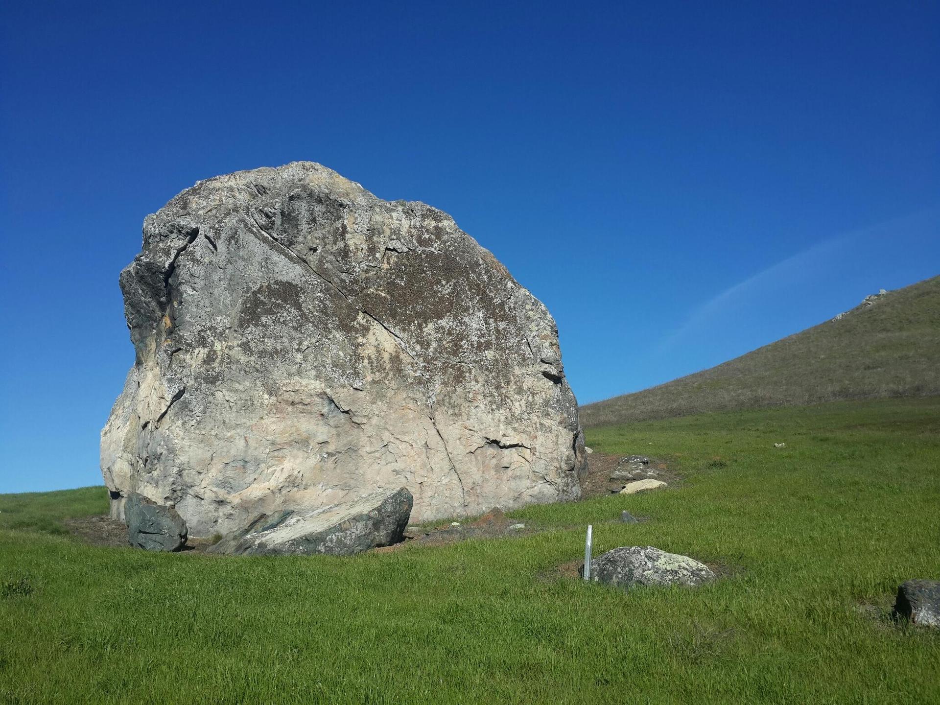 A large boulder sits on a bright green grassy hill under a clear blue sky.