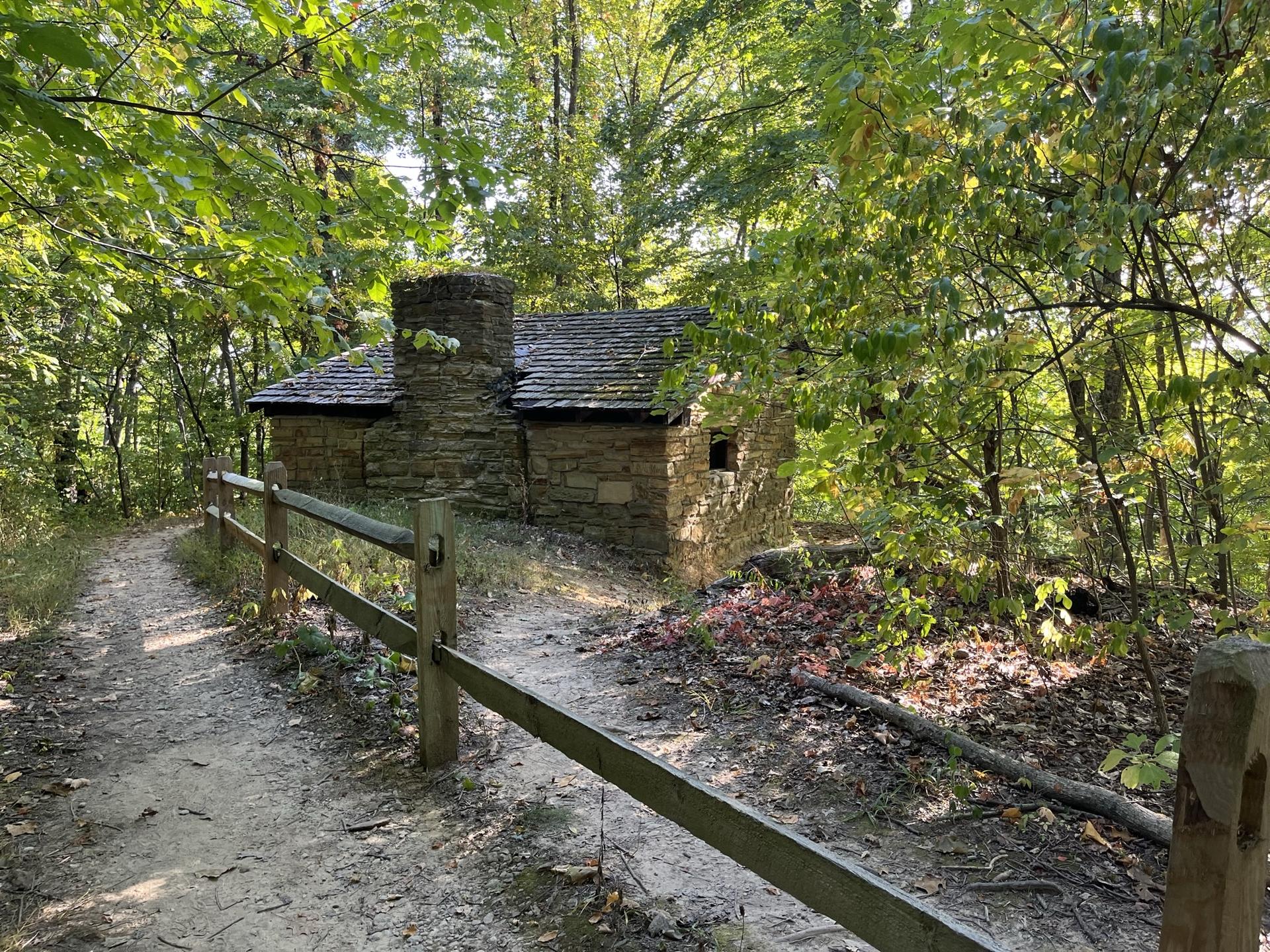 Small stone building with chimney by a dirt path in a dense green forest.