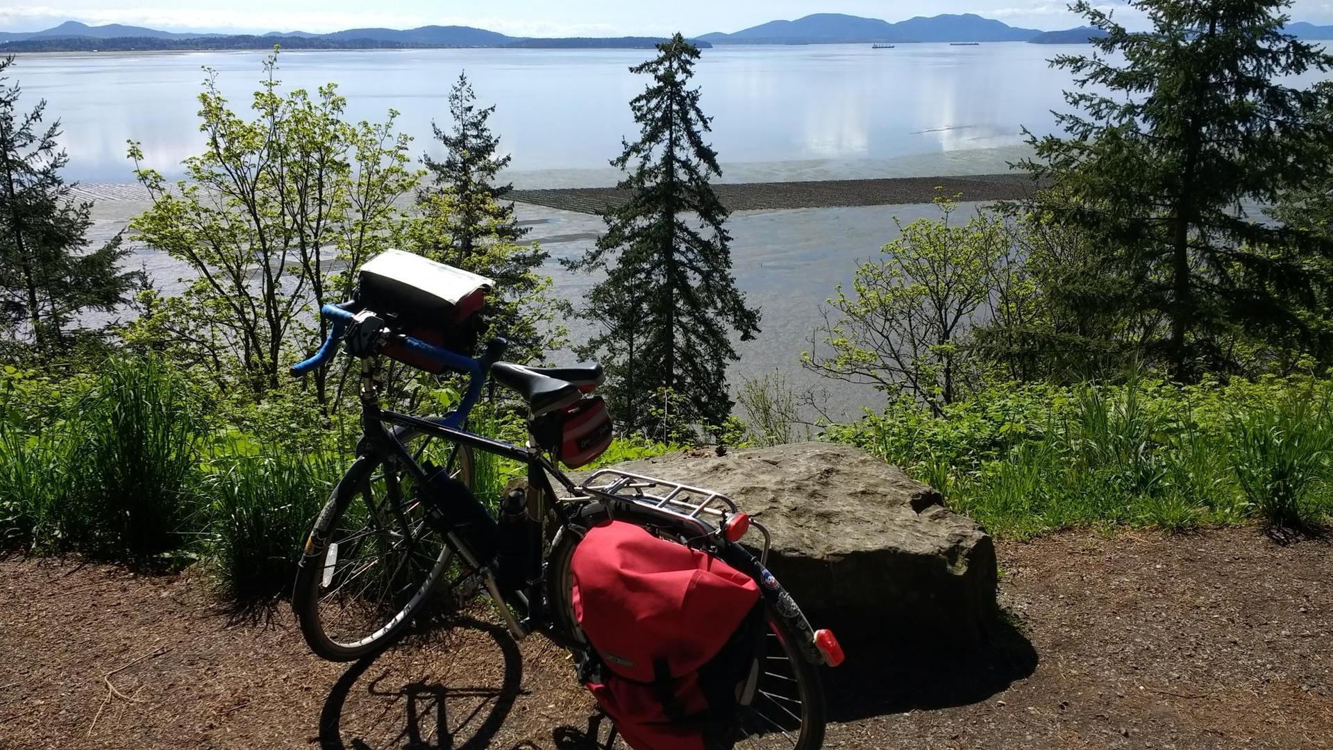 Bicycle with red bags parked overlooking a sunny bay with distant mountains.