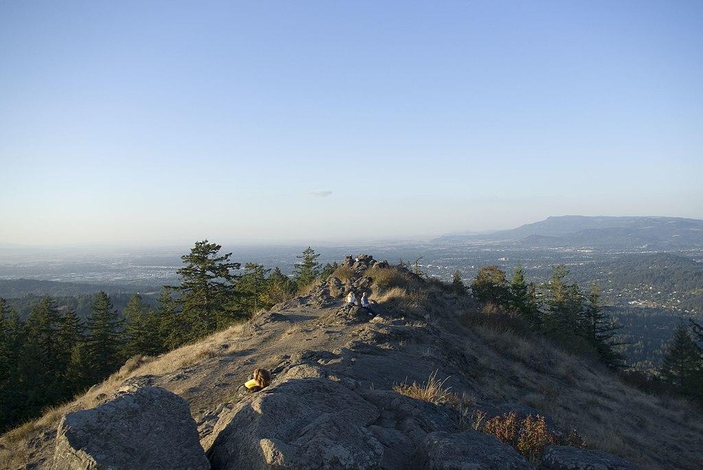 People gather on a rocky mountain ridge overlooking a distant city at sunset.