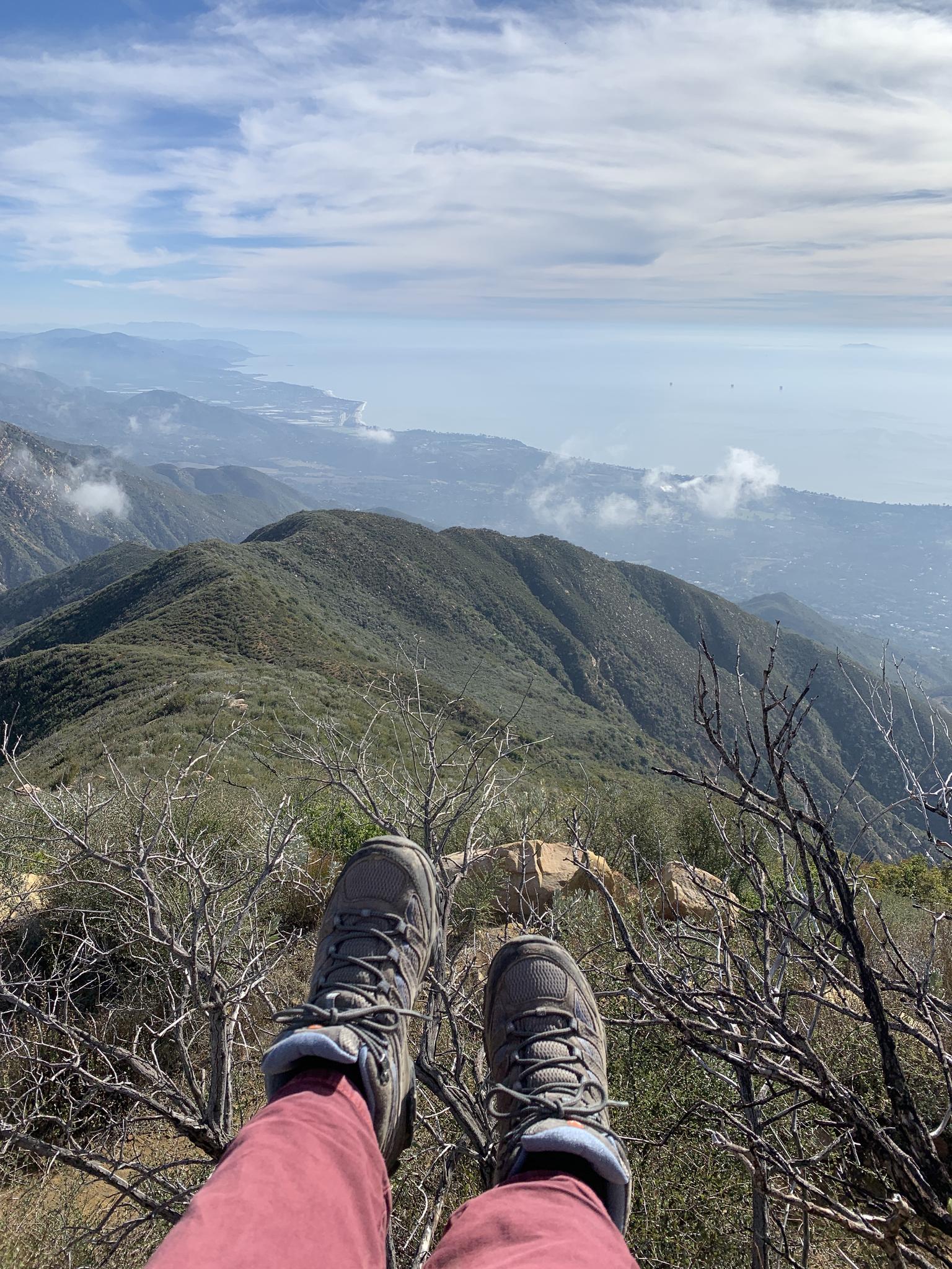 Hiking boots and legs dangle over a vast mountain and ocean vista.