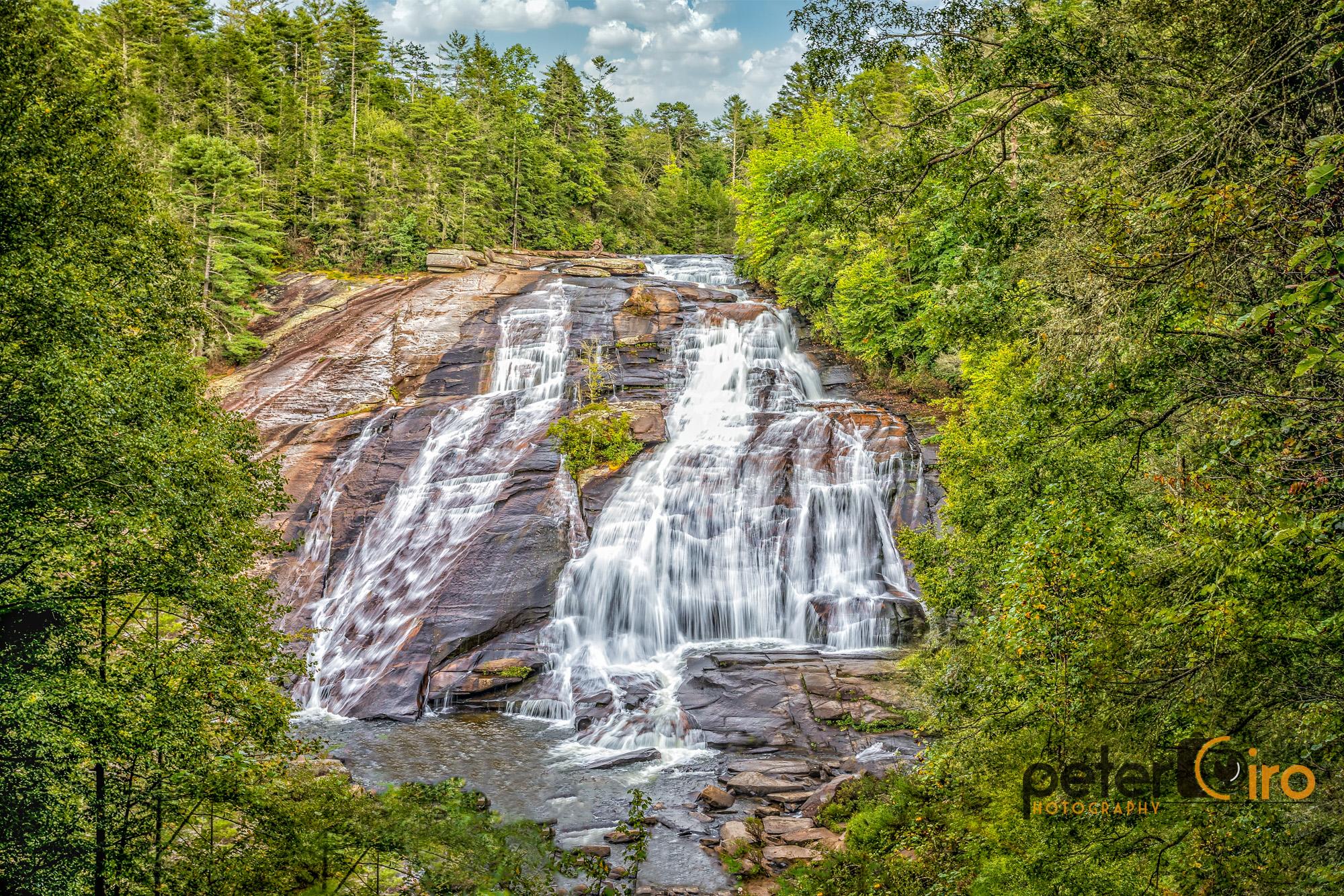 Wide waterfall flows over dark rocks, surrounded by lush green forest.