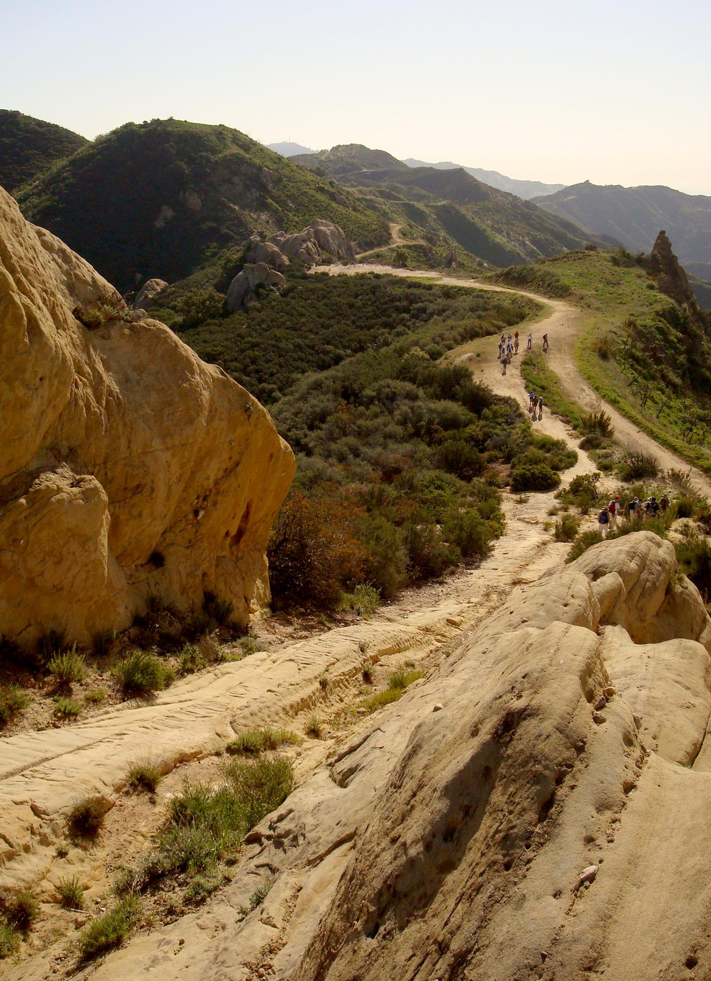 Hikers walk a winding dirt path on a rocky, green hillside in bright sun.