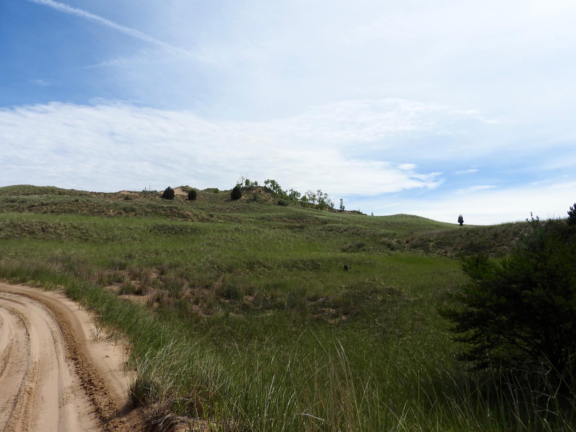Sandy track winding beside lush green grassy hills under a partly cloudy sky.