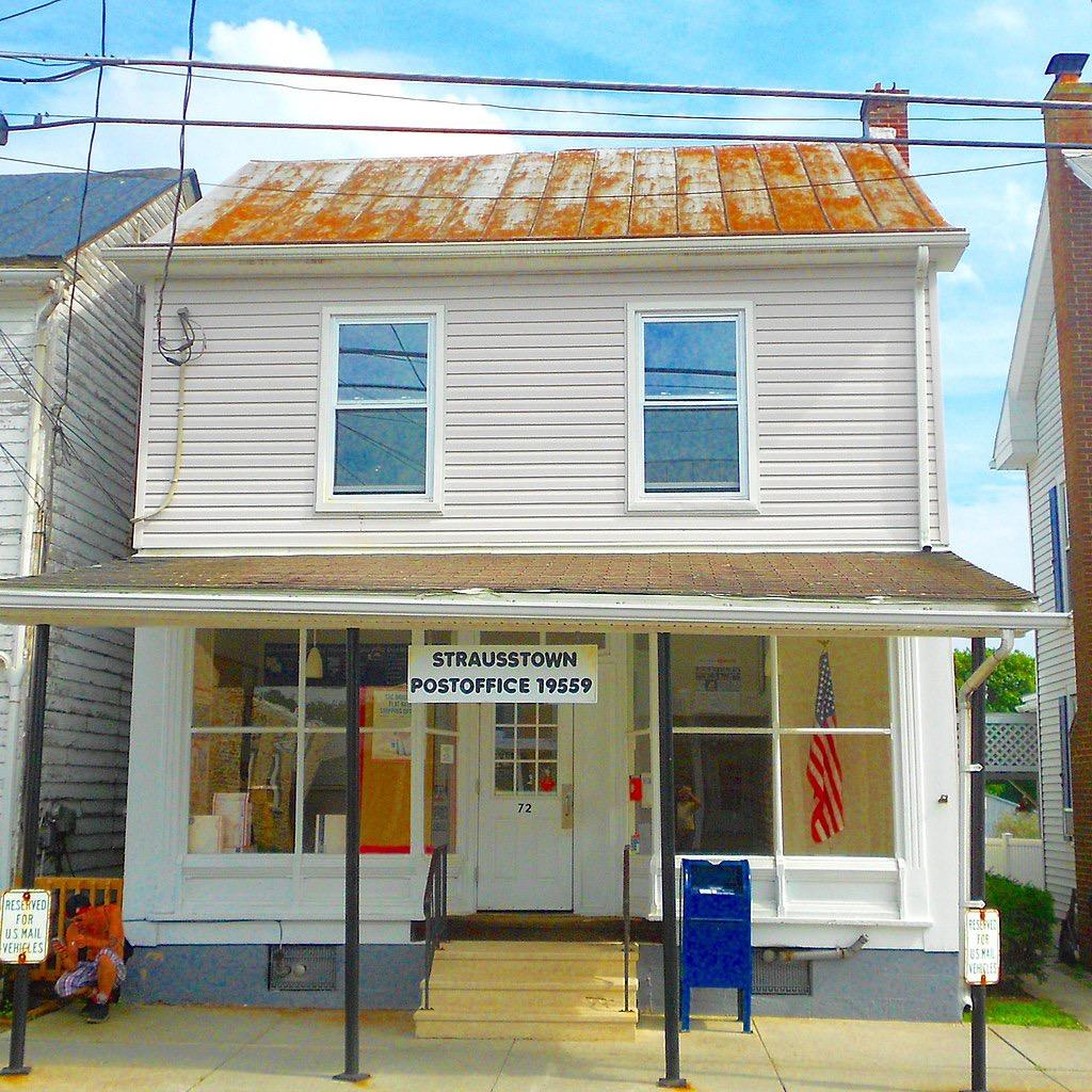 A light-colored building with a rusty roof, porch, and blue mailbox in daylight.