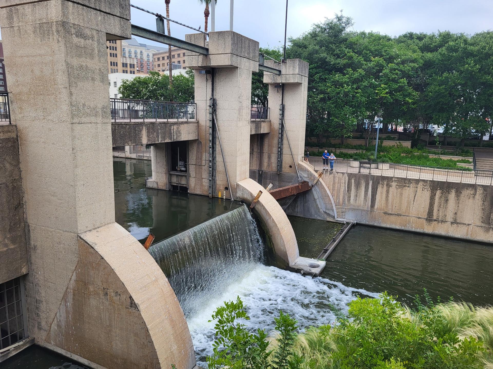 Concrete dam with water cascading, two people on a walkway, green trees.