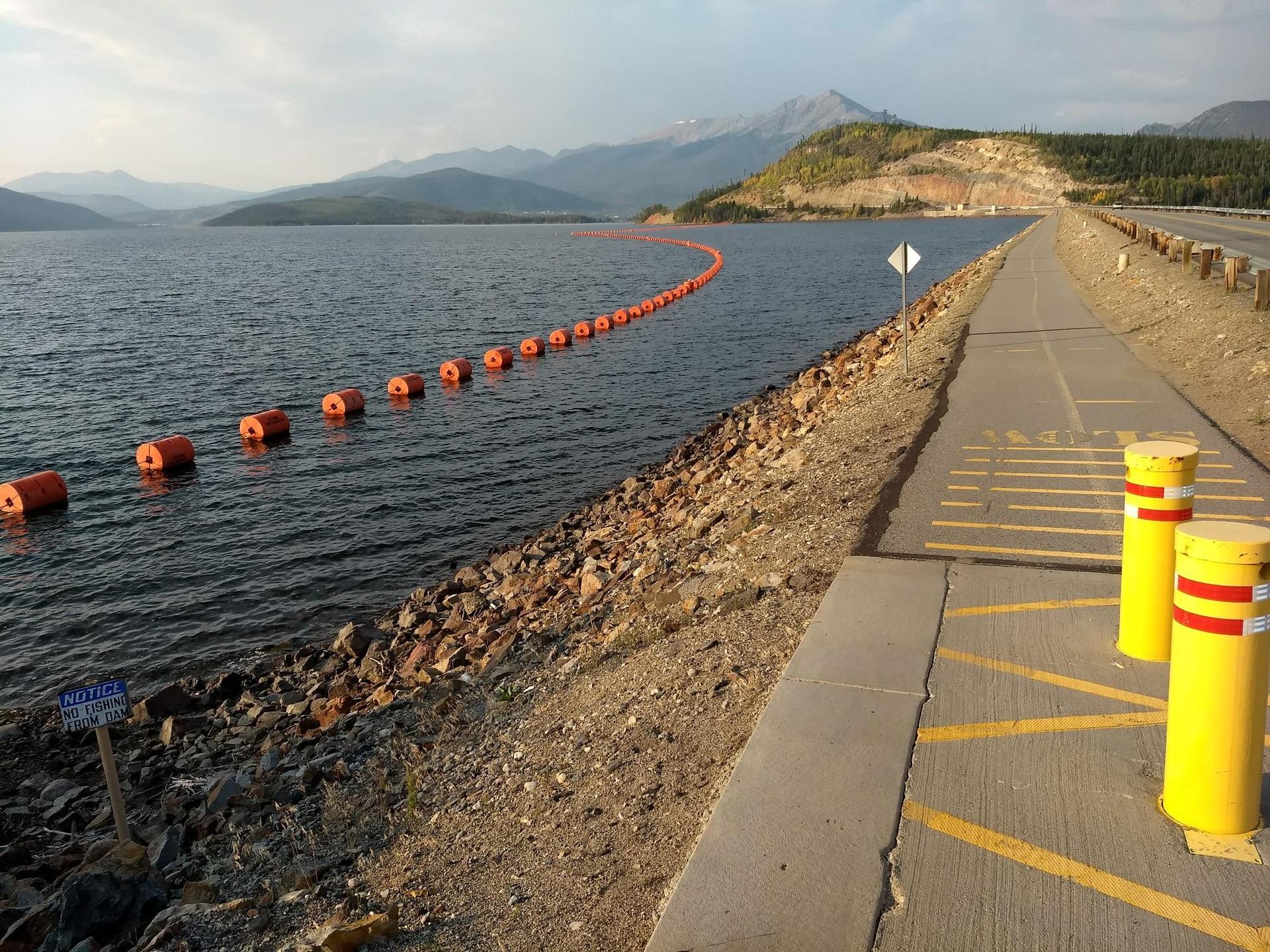 Paved road beside a lake with orange buoys and distant mountains.