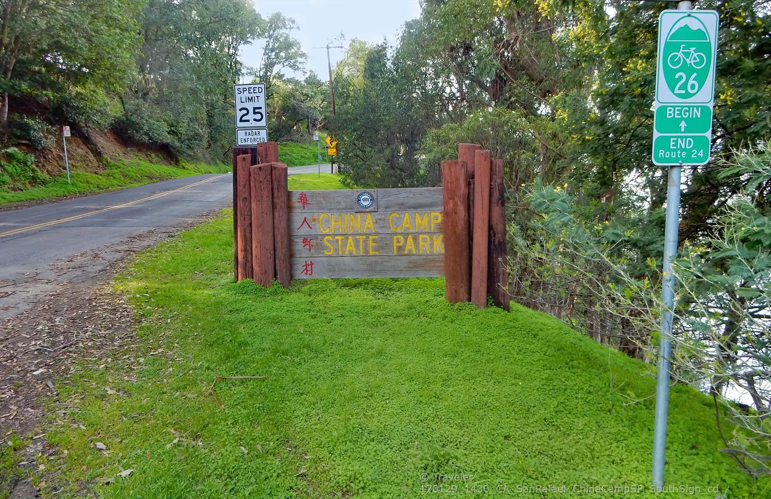A wooden sign and a bike route sign stand on a grassy slope by a road.