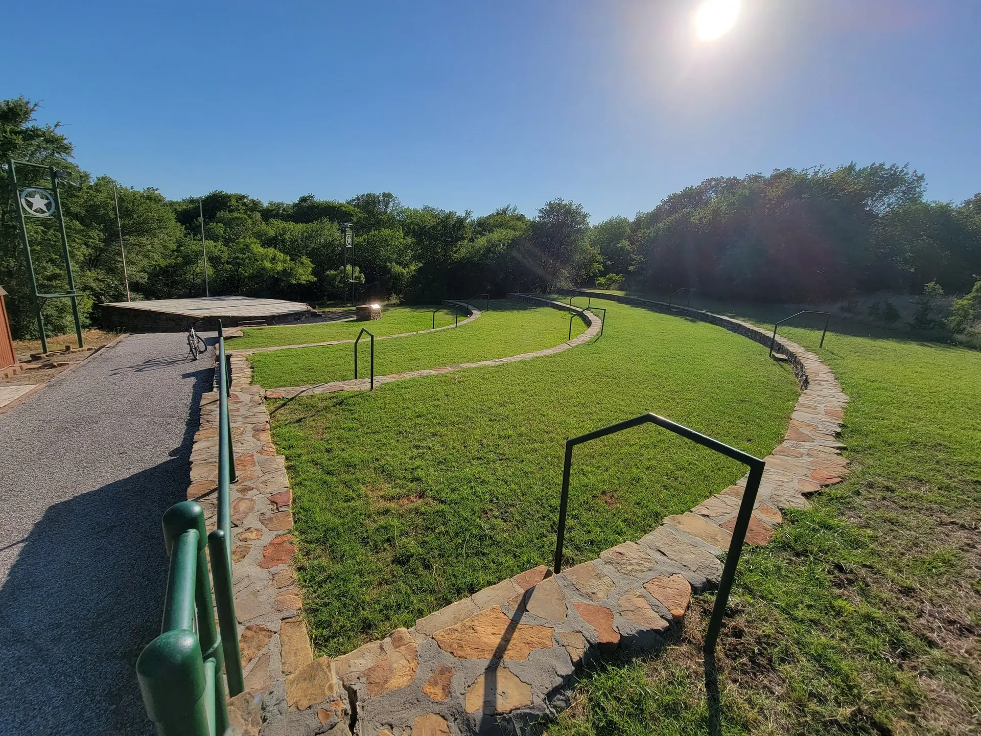 Sunny outdoor amphitheater with green grass seating and a stage.