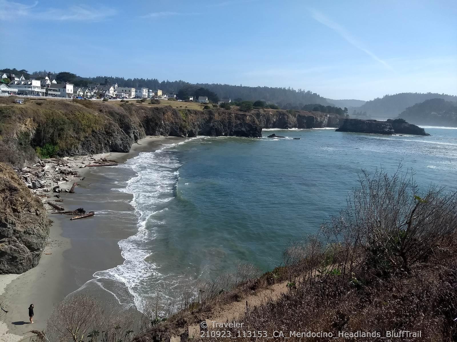 Sunny coastal inlet with sandy beach, cliffs, distant town, and one person.