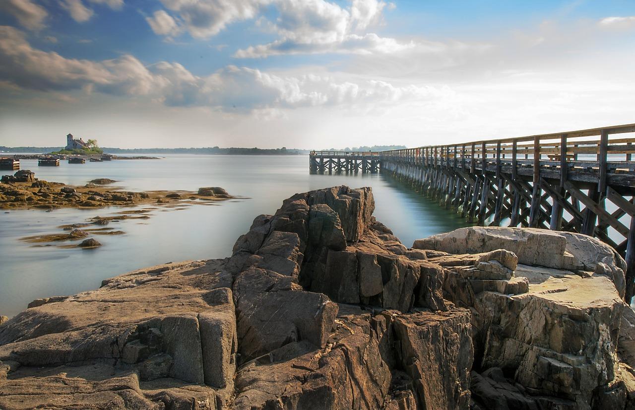 Sunny coastal view with large rocks, a long pier, and distant island.