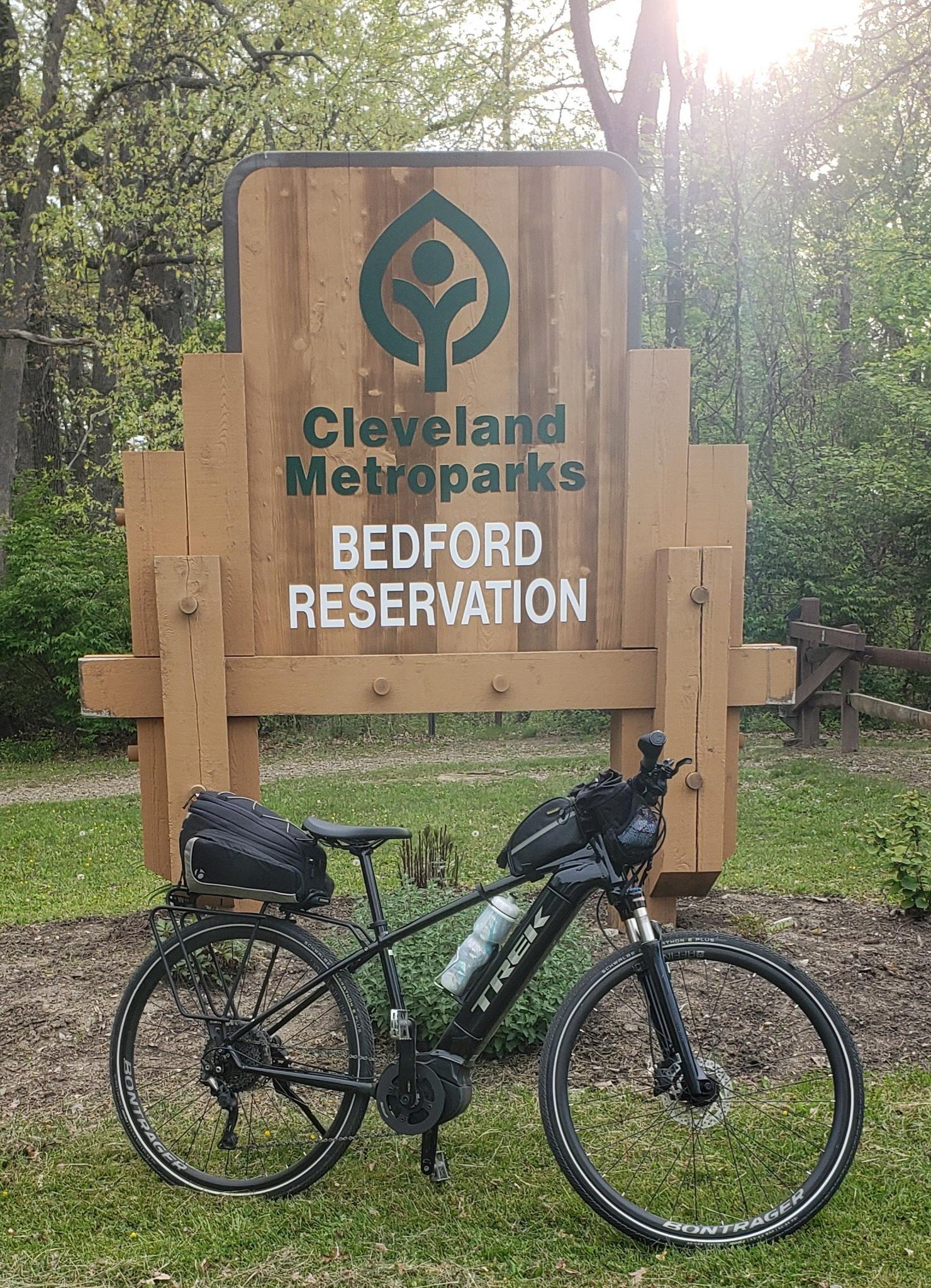 A black bicycle parked in front of a large wooden sign in a sunny forest.