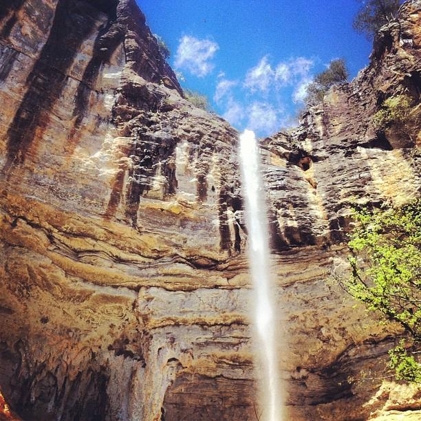 A tall waterfall cascades down steep rock cliffs under a bright blue sky.
