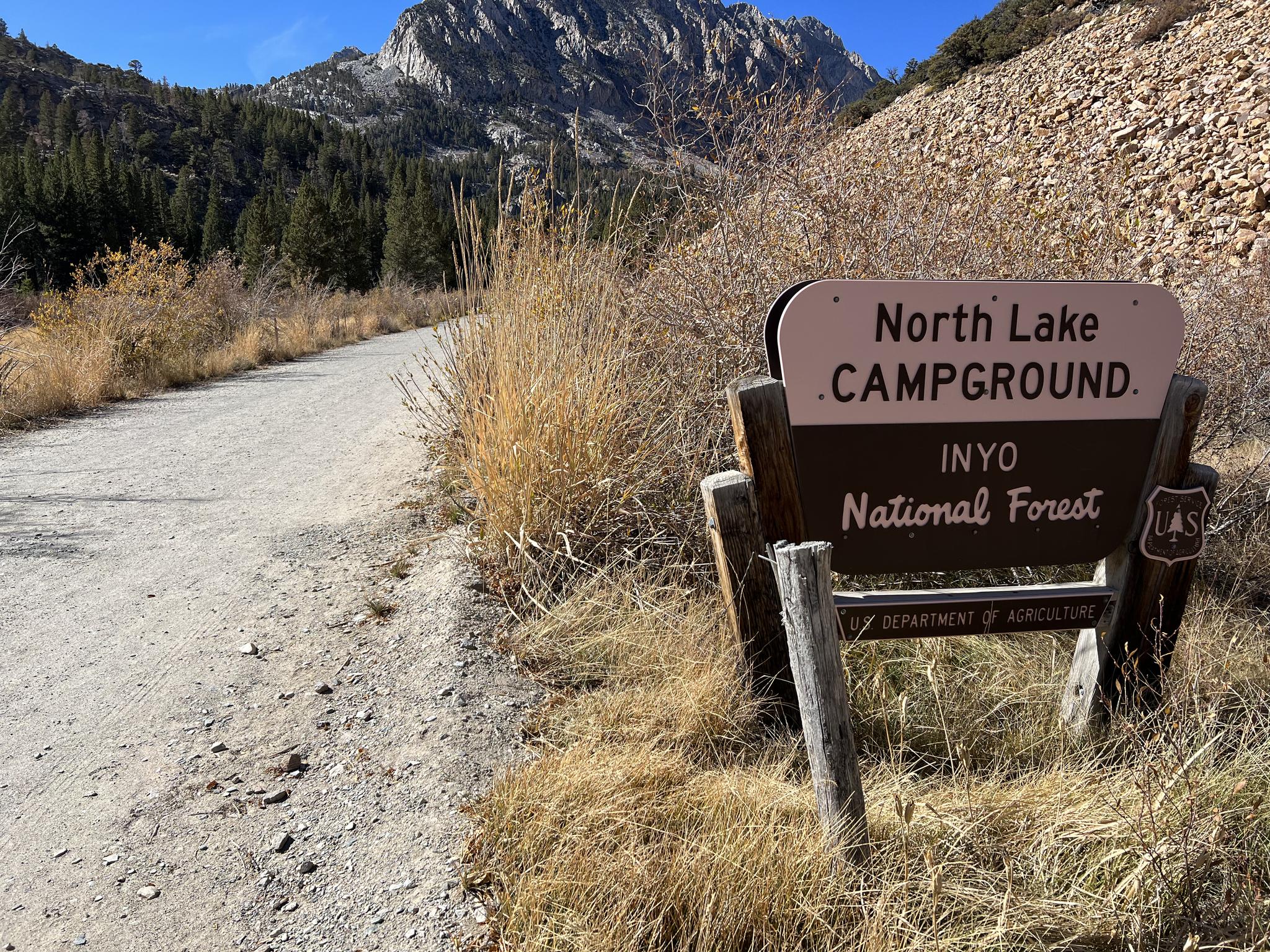 A campground sign on a dirt road with mountains and trees under a clear sky.