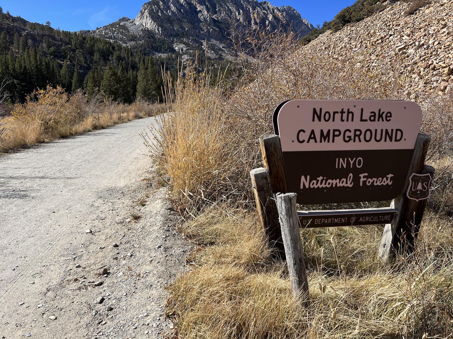A campground sign on a dirt road with mountains and trees under a clear sky.