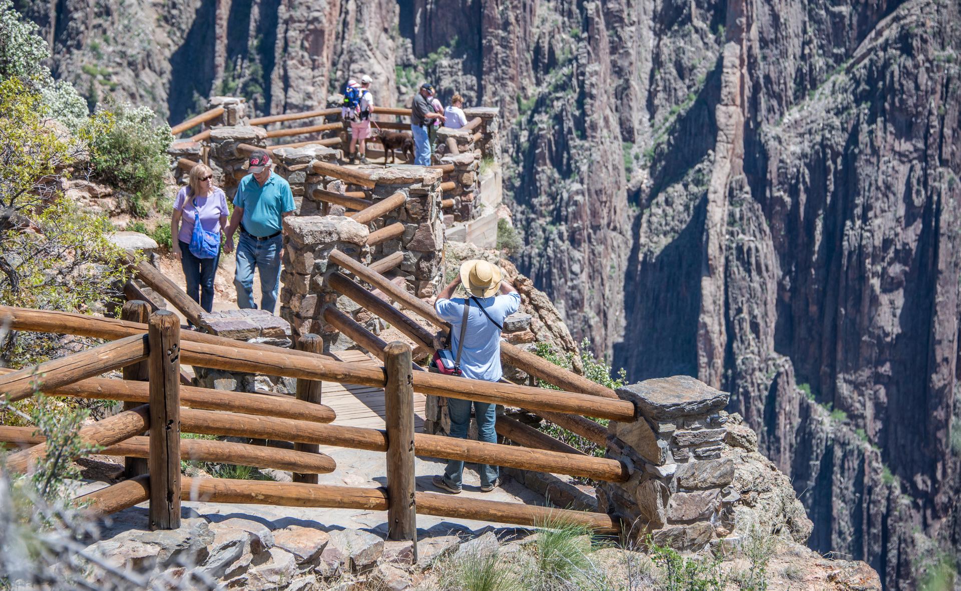 People walk on a wooden railing path along a sunny, rocky canyon.