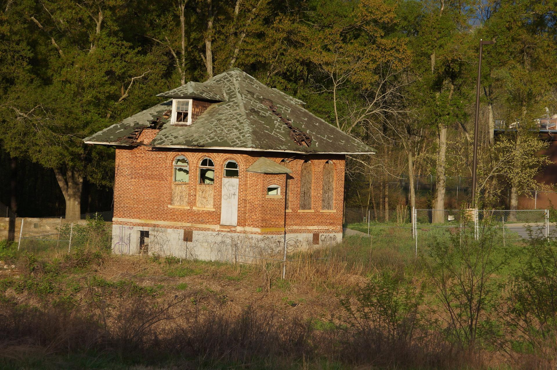 An old brick building with a green roof stands among trees in sunlight.