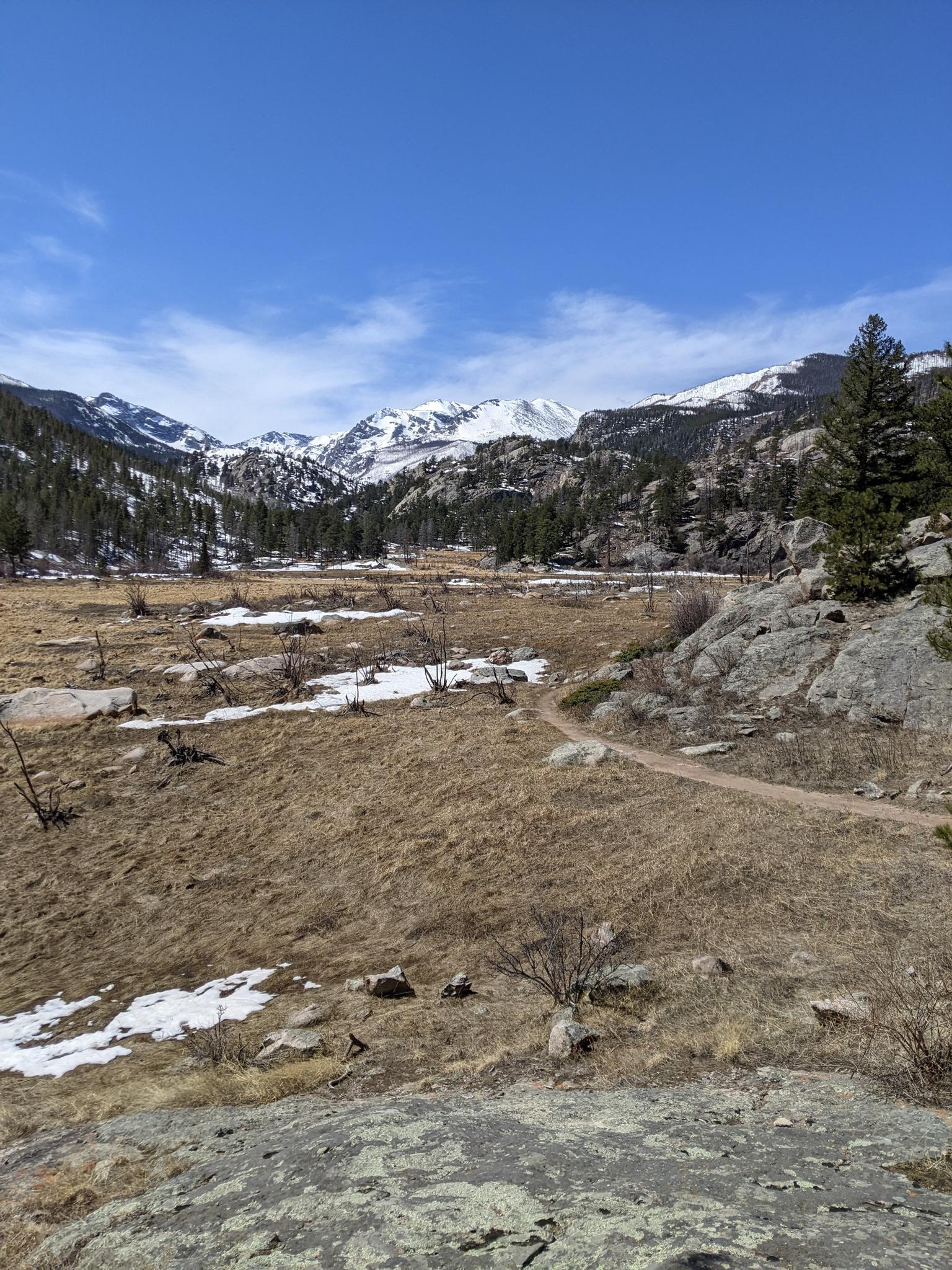 A dirt path winds through a dry valley with snow-capped mountains.