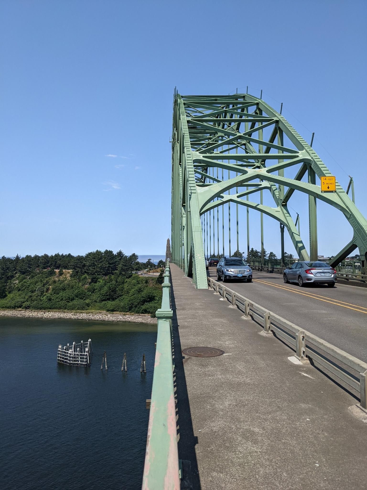 Cars drive on a green arched bridge over water on a sunny day. - Yaquina River loop