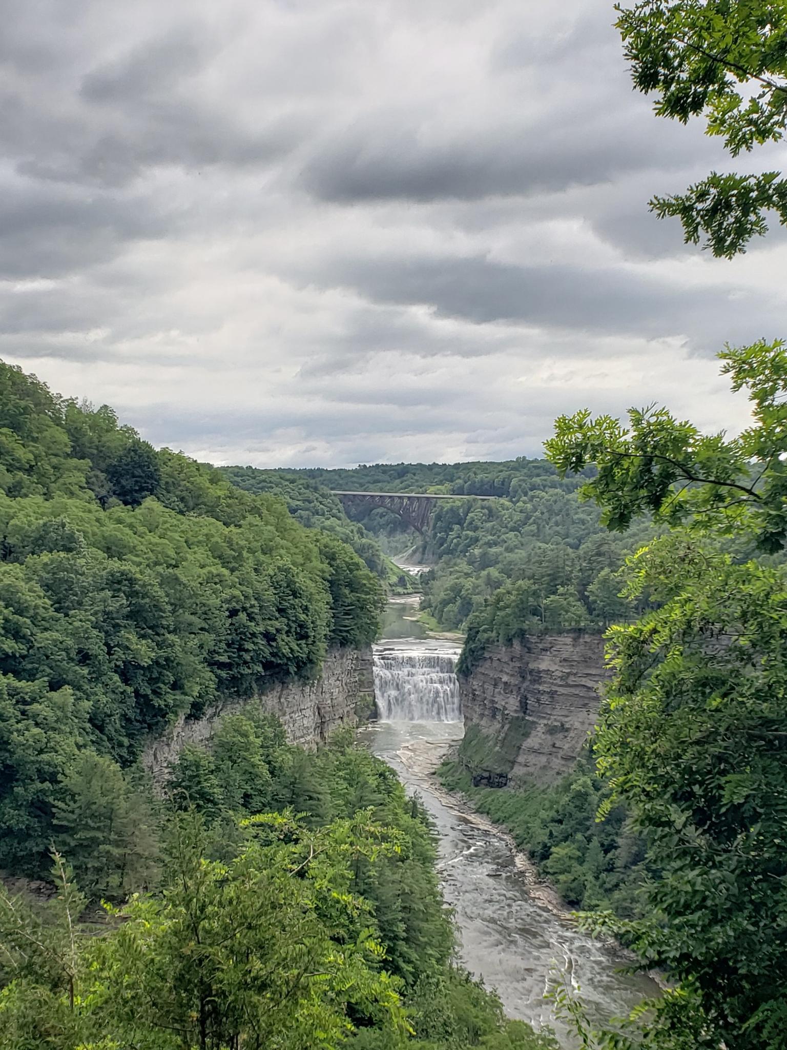 Waterfall in a deep gorge with a distant bridge under cloudy skies.