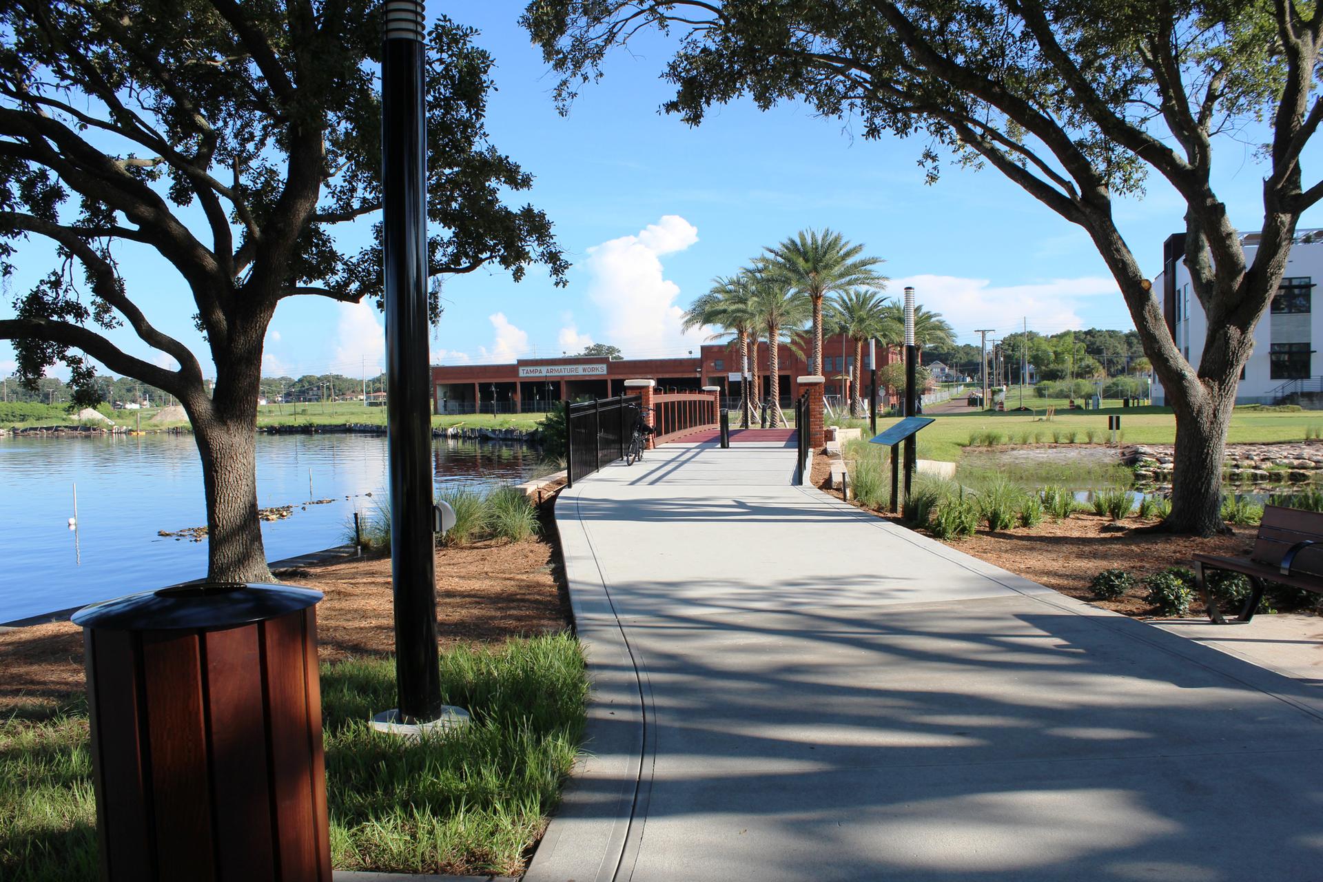 Paved walkway along water, shaded by trees, leading to a building.