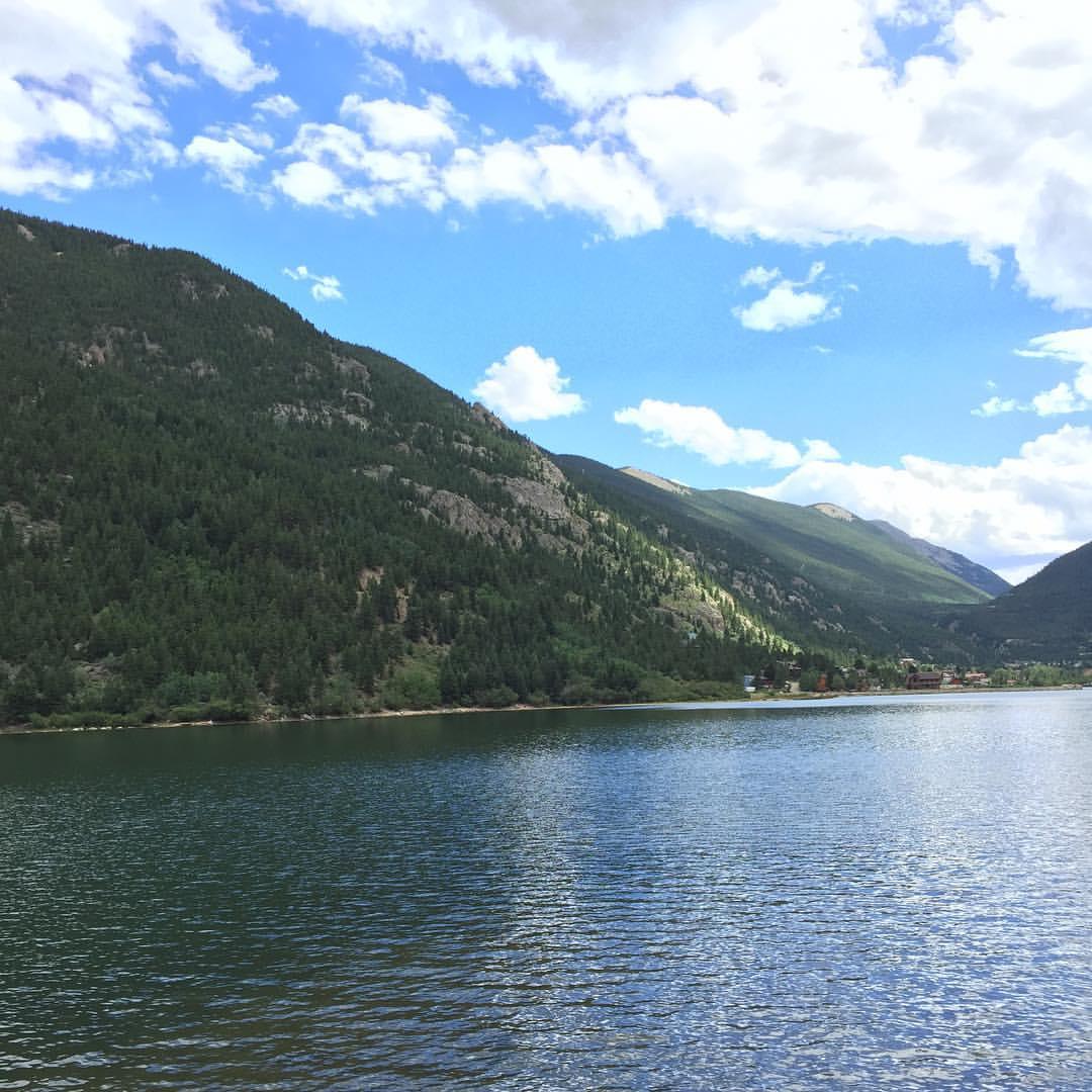 Wide lake with green mountains and distant buildings under a partly cloudy sky.