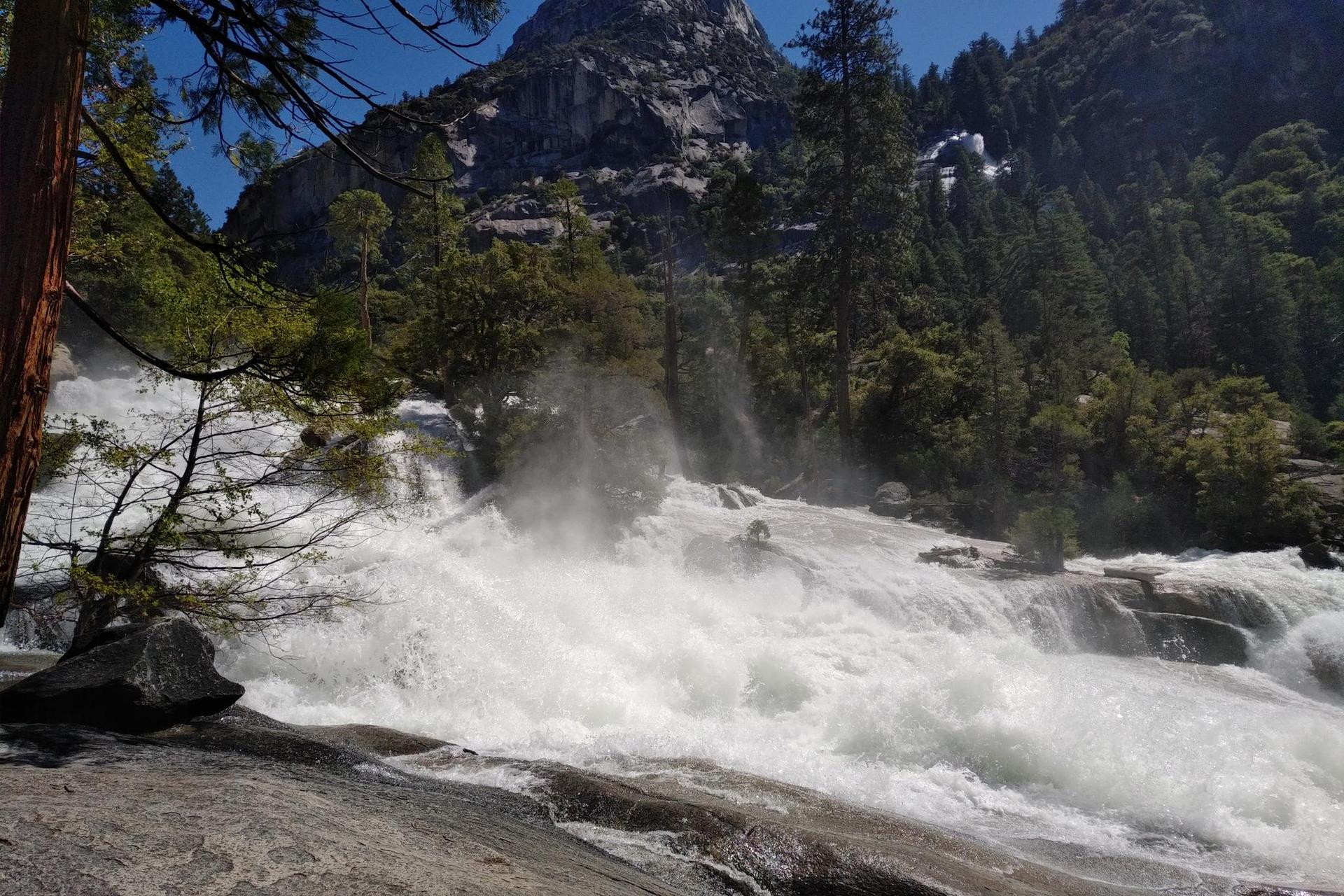 Powerful white water rapids crash over rocks in a sunny mountain landscape.
