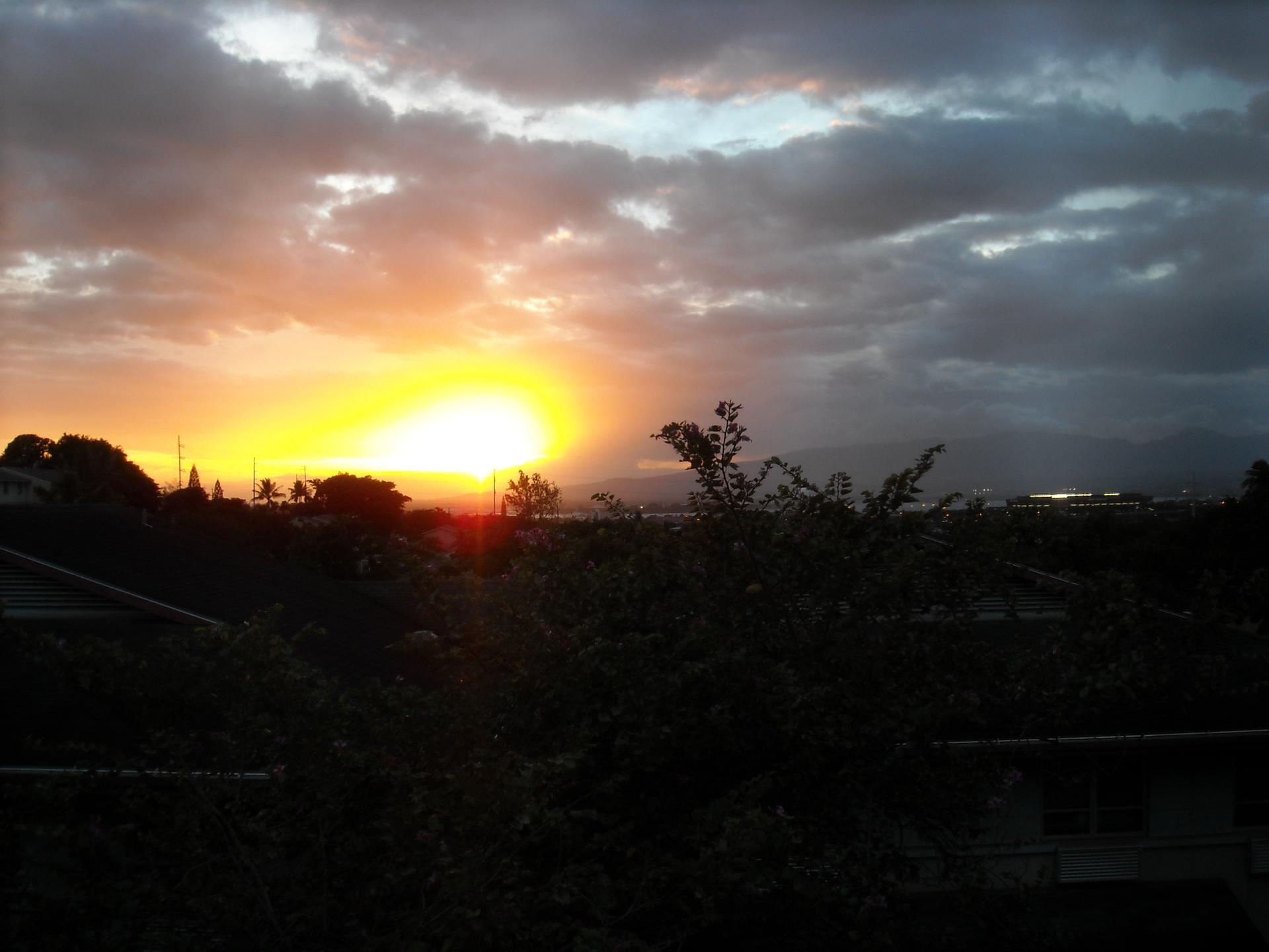 Golden sunset over a dark, silhouetted landscape with cloudy skies and distant lights.