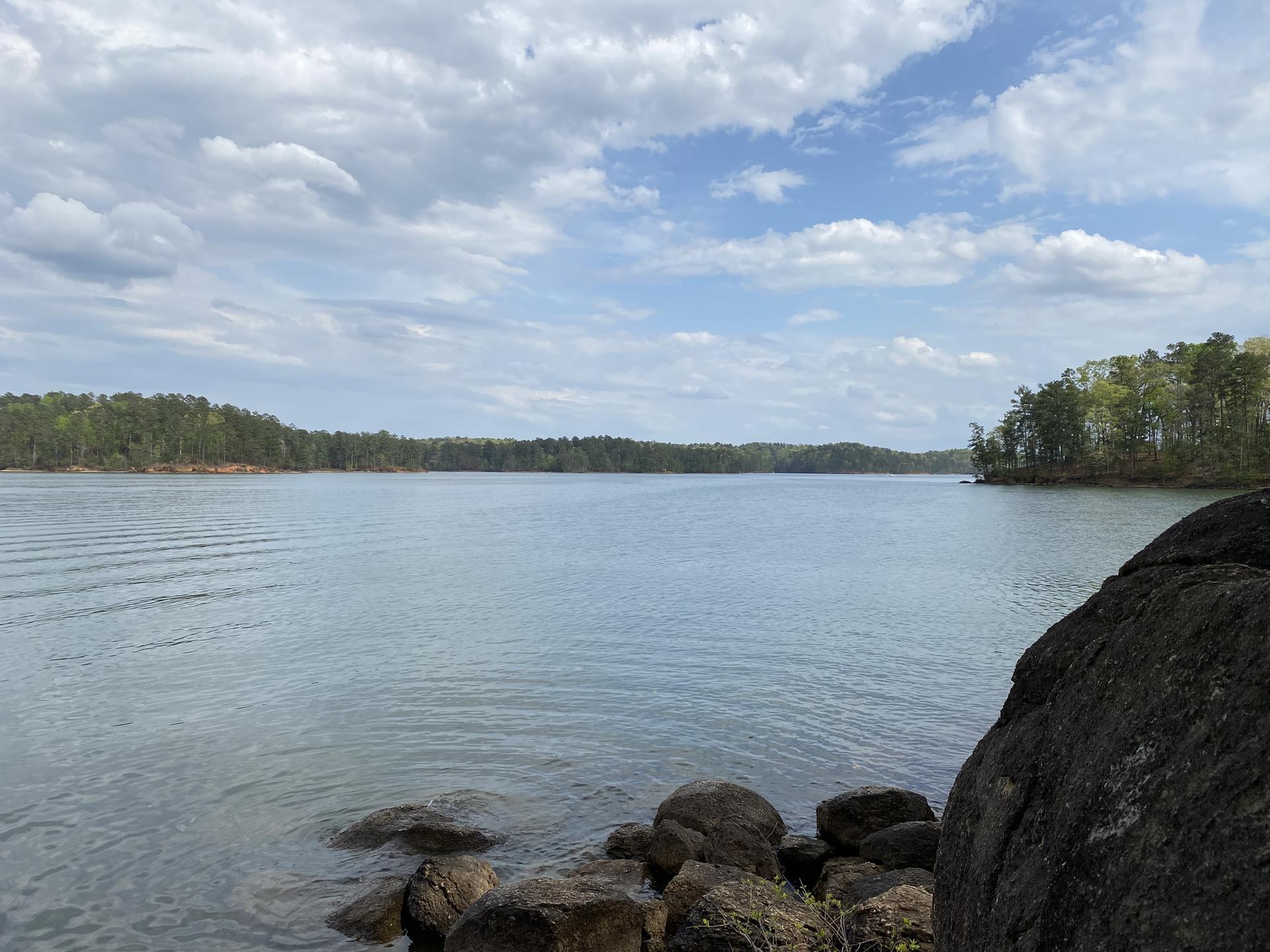 Wide view of a large lake surrounded by green forests under a cloudy sky.