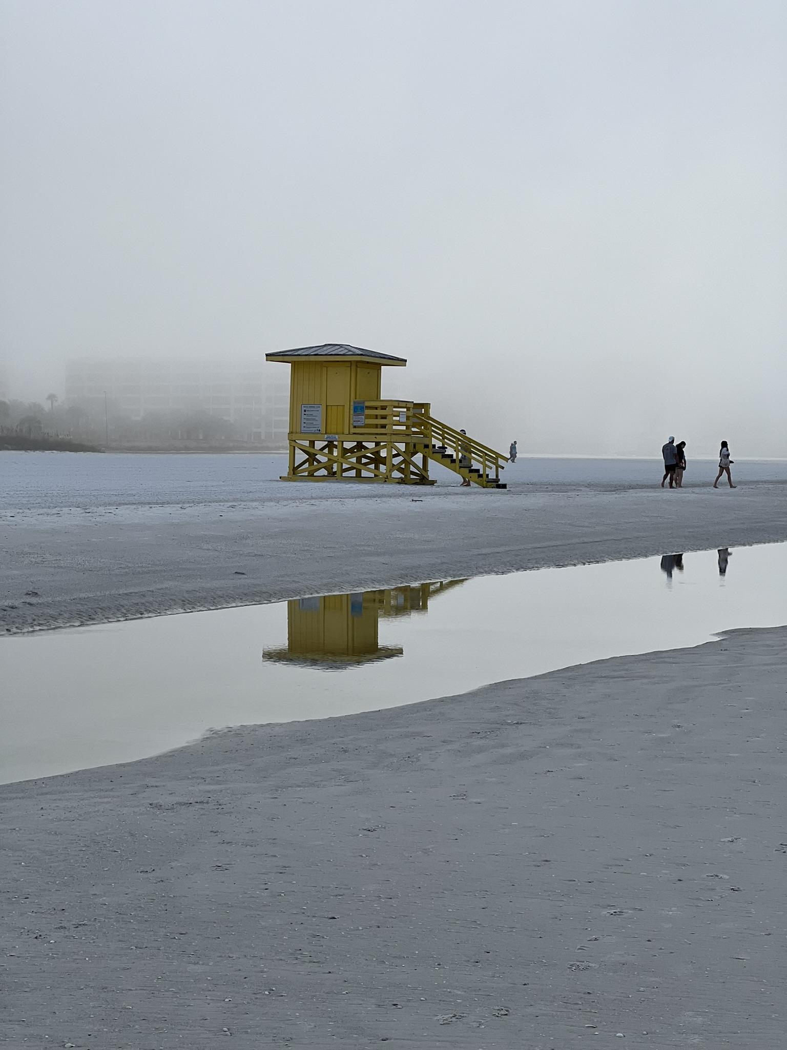 Yellow lifeguard stand on a foggy beach, reflected in a shallow pool.