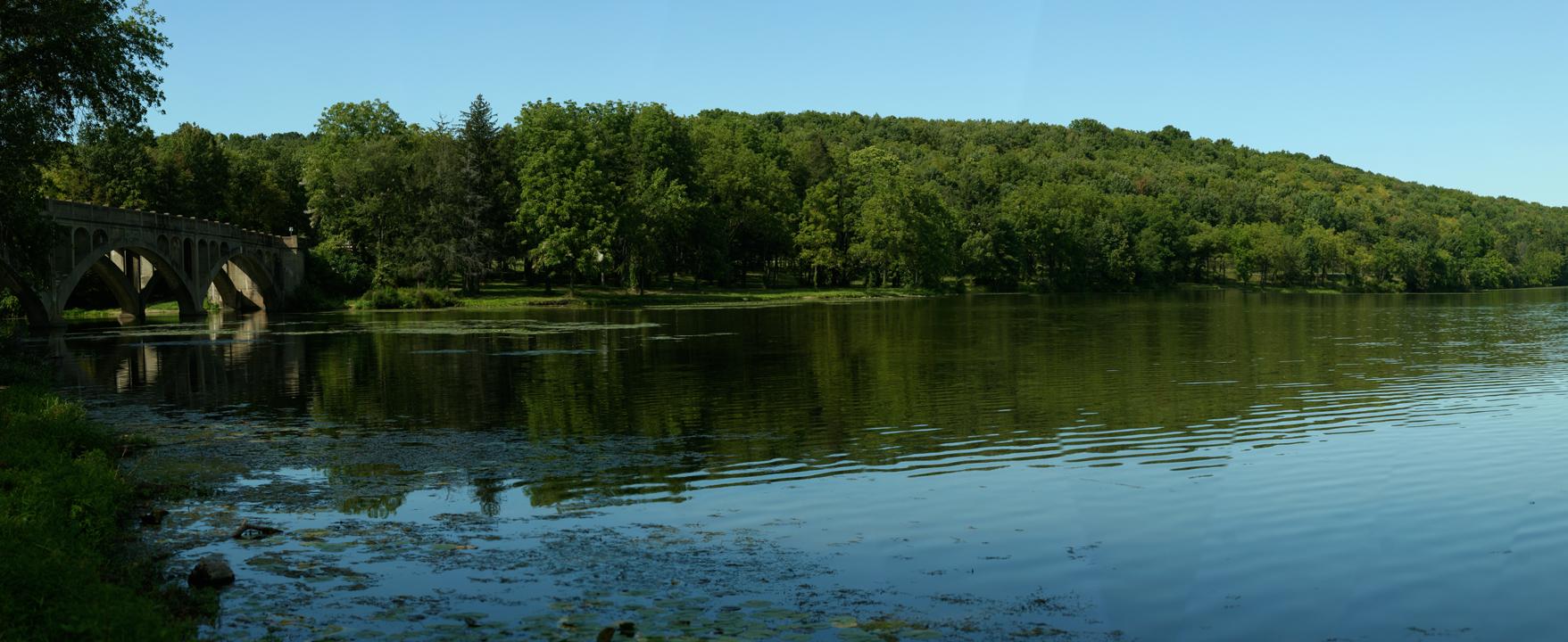 Stone bridge over calm water, surrounded by lush green trees under a clear sky.