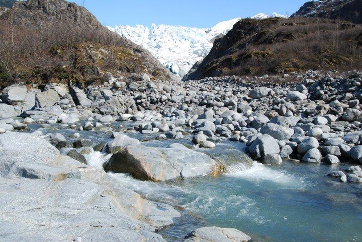 Rocky river flows through a valley with snow-capped mountains under clear skies.