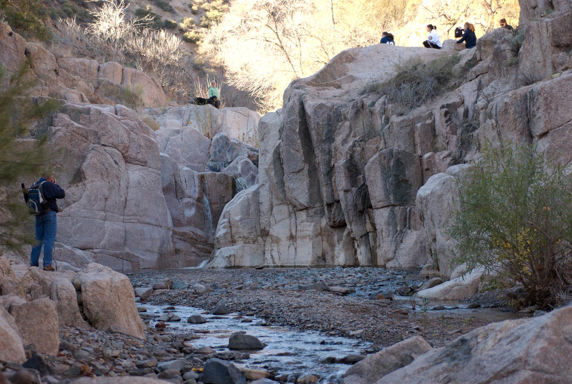 A person stands by a rocky stream in a sunlit canyon with others on cliffs.