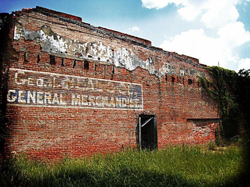 Weathered brick building with a faded "General Merchandise" sign and overgrown grass.
