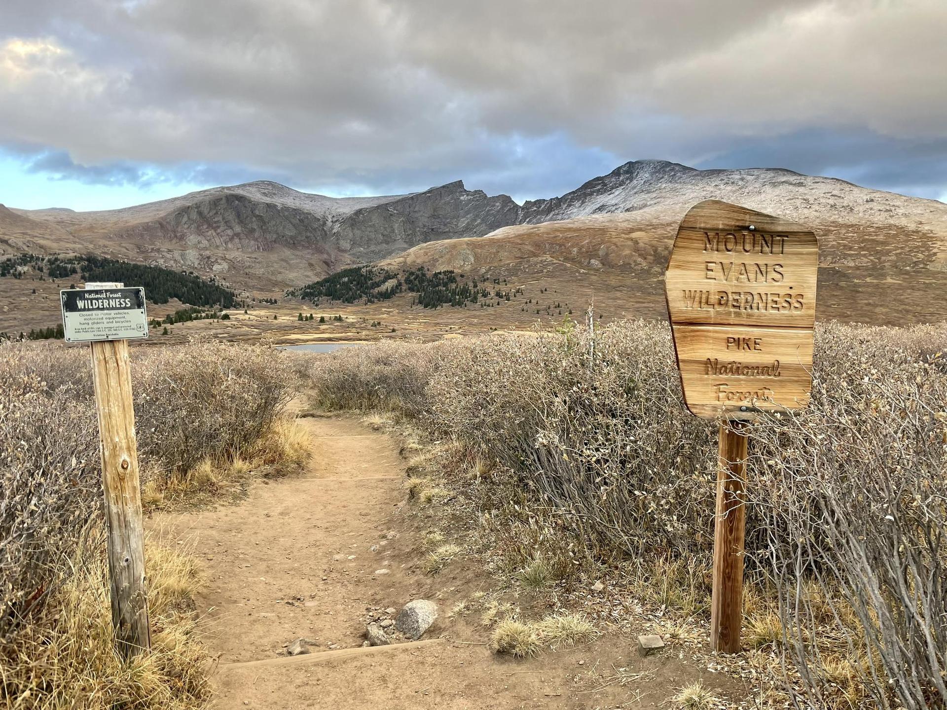 Dirt path leads to distant mountains under cloudy sky, with wooden trail signs.