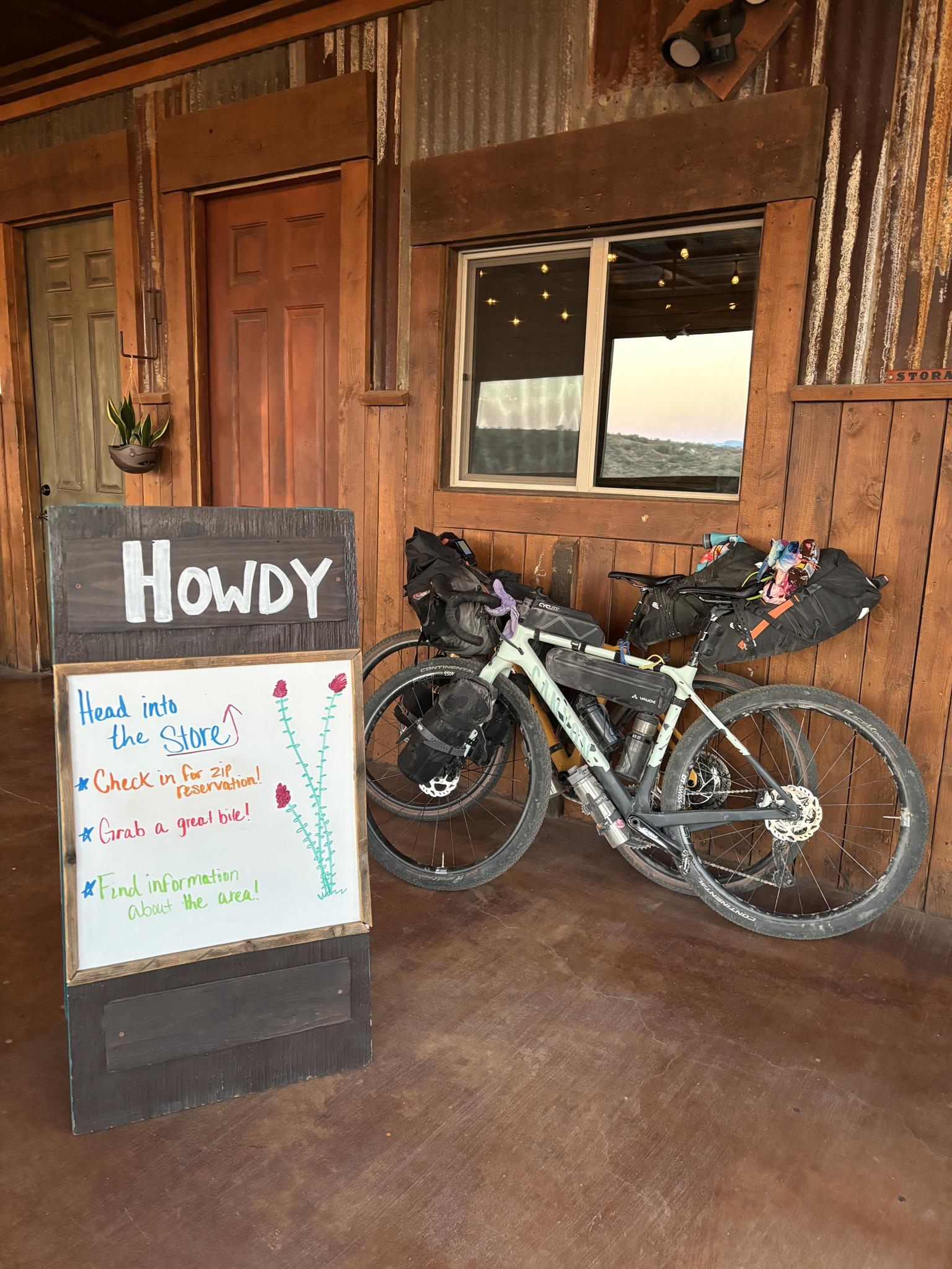 Loaded bicycle parked next to a "HOWDY" sign on a wooden porch.