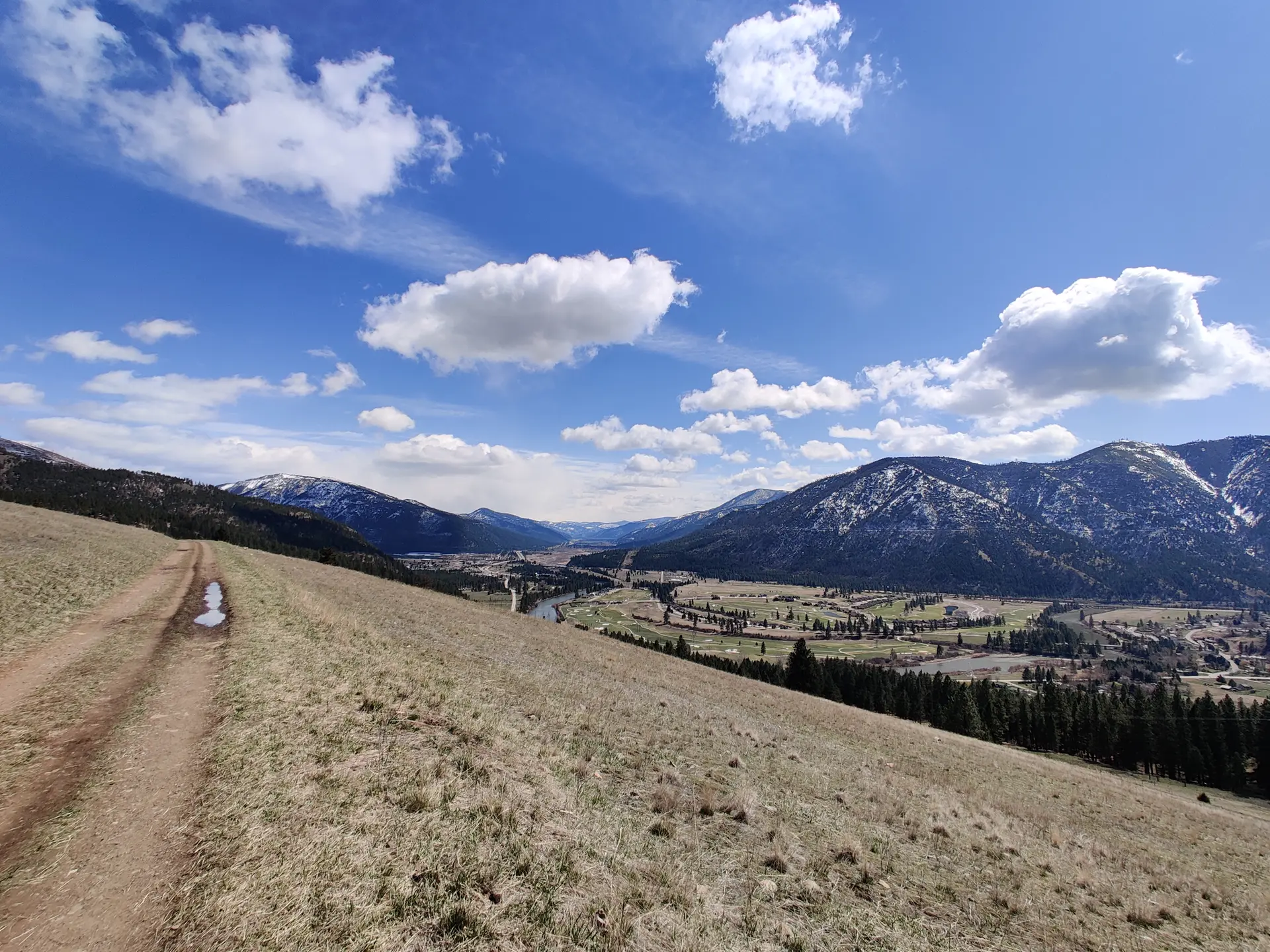 Dirt path on grassy hillside overlooking a valley with snow-capped mountains under a bright sky.