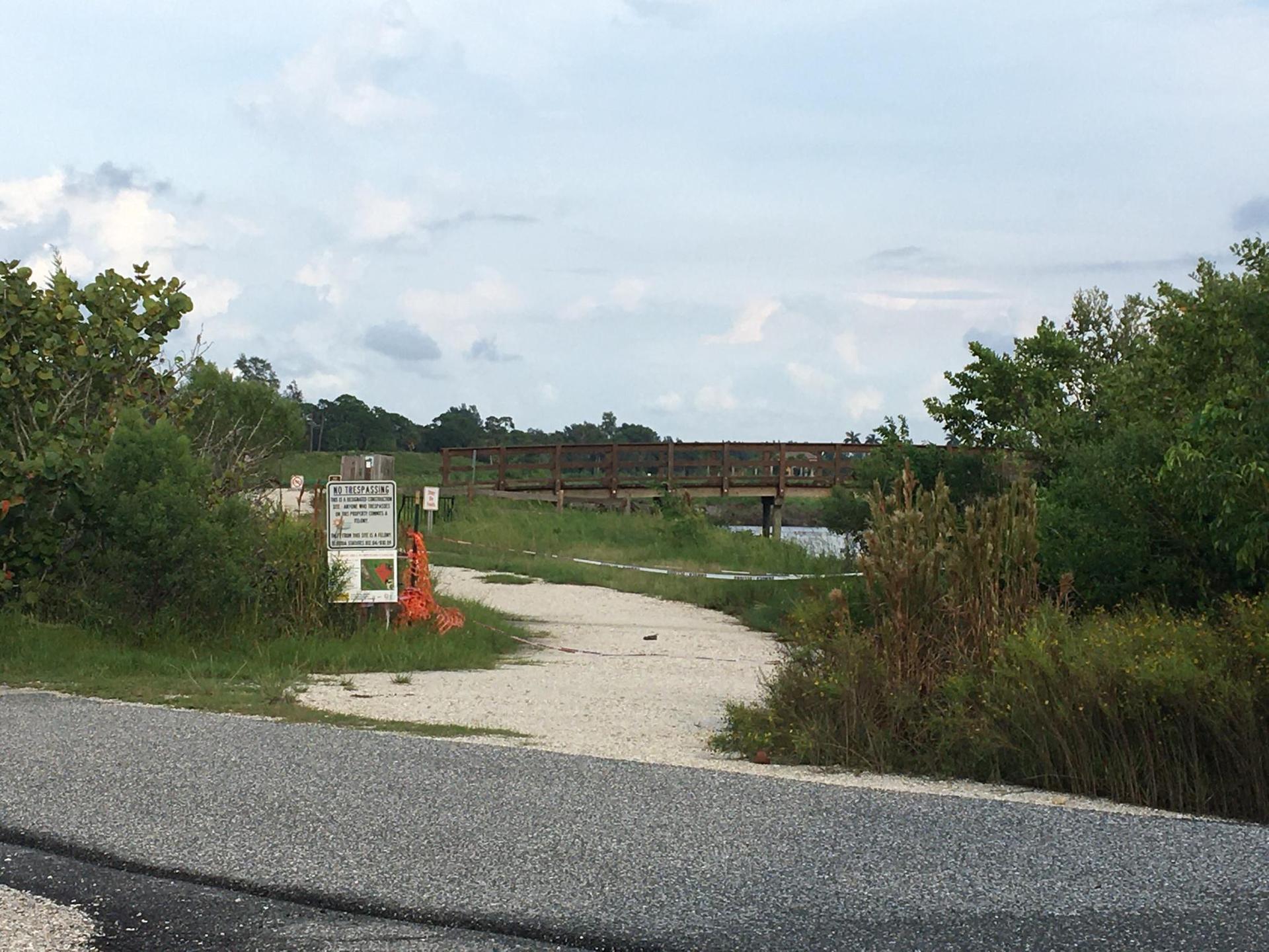 A wooden bridge spans a waterway, seen from a gravel path under a cloudy sky.