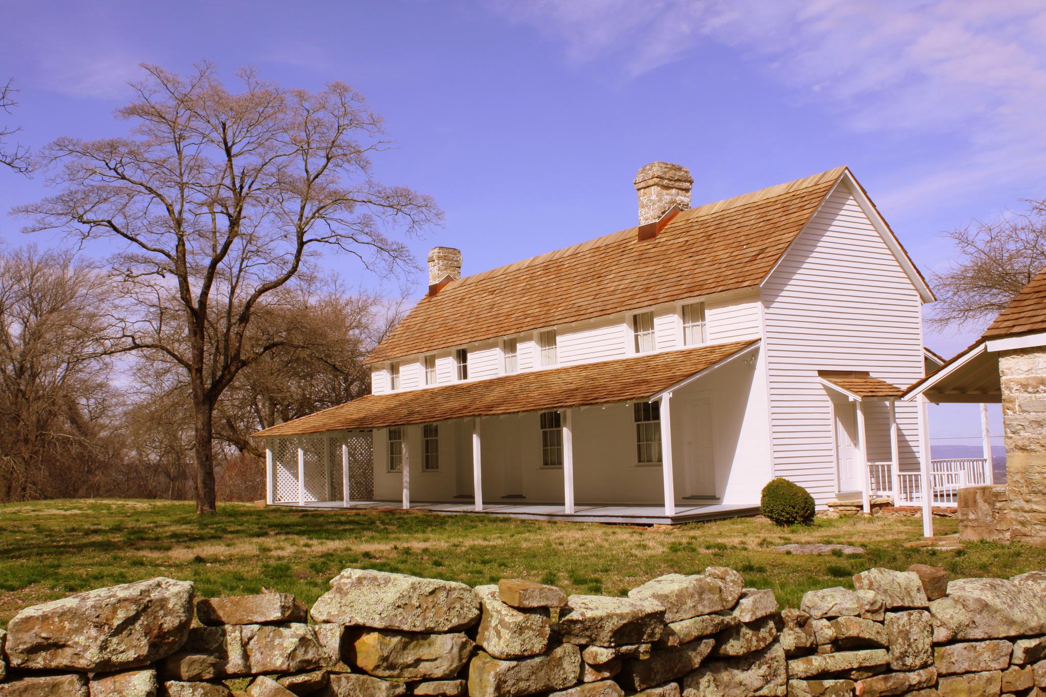 White historic house with a long porch and stone wall under blue sky.