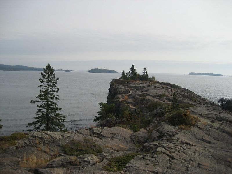Rocky cliff with pine trees overlooks a vast body of water and islands.