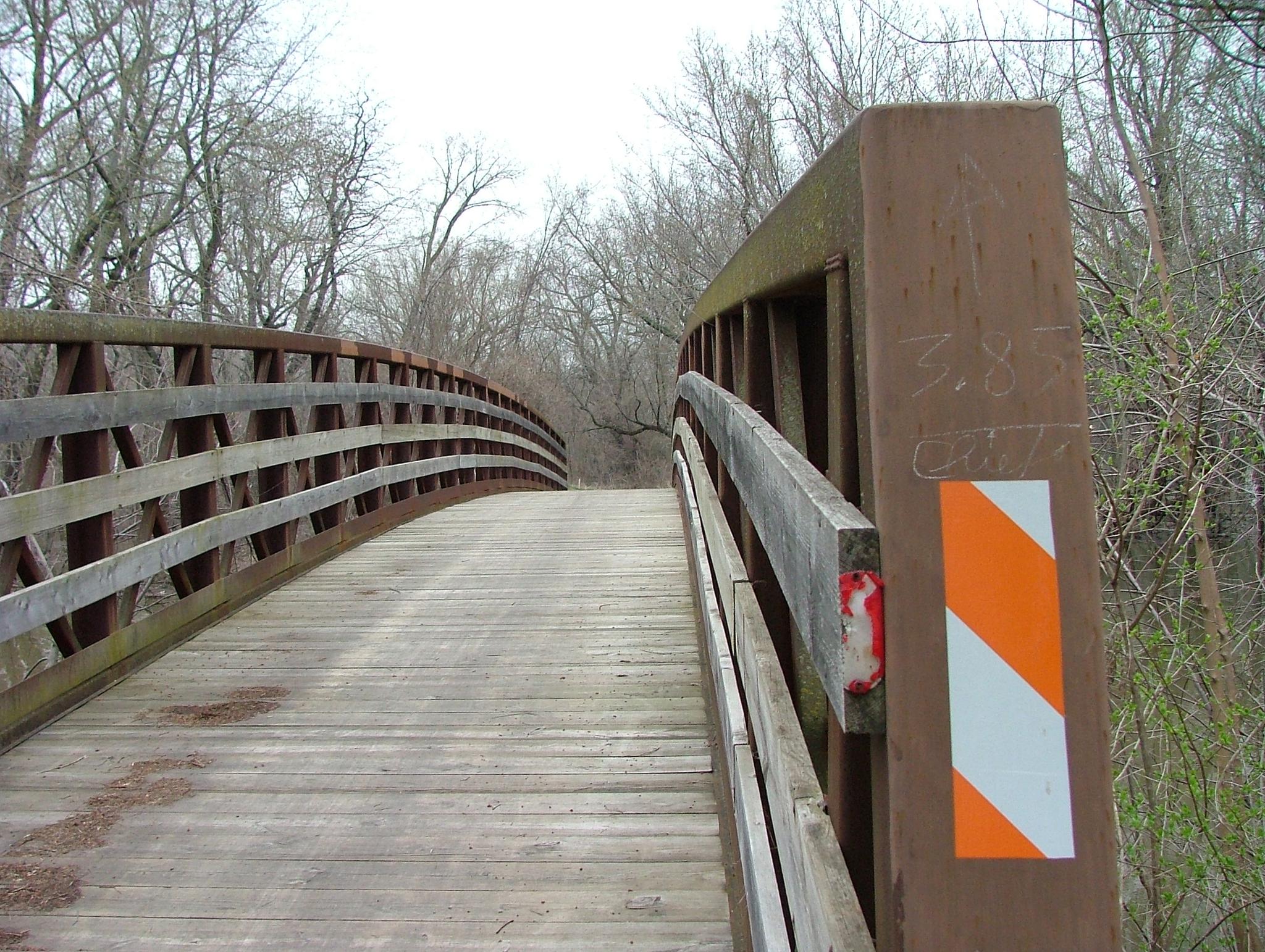 Curved wooden bridge with railings, leading into a bare winter forest.