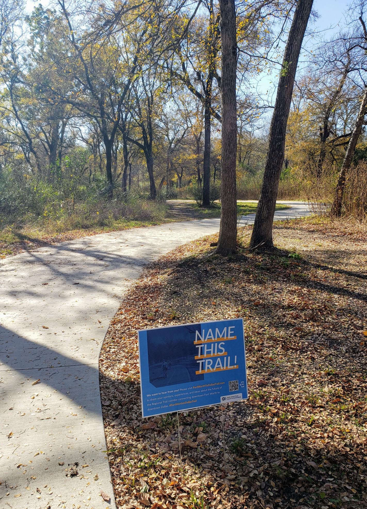 Sunny paved path winding through autumn woods, with a blue sign.
