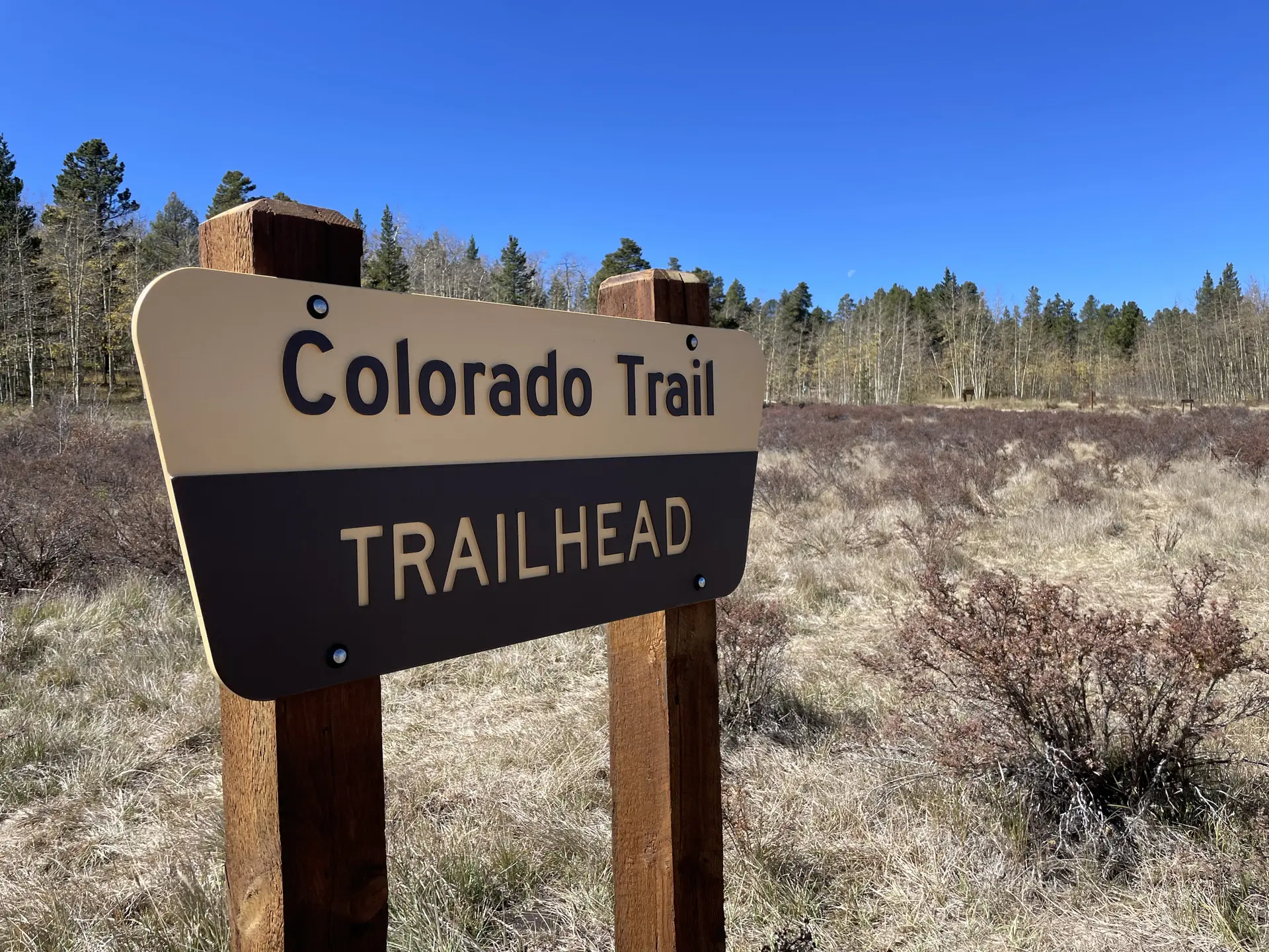 A trailhead sign in a dry grassy field with a forest under a clear sky.