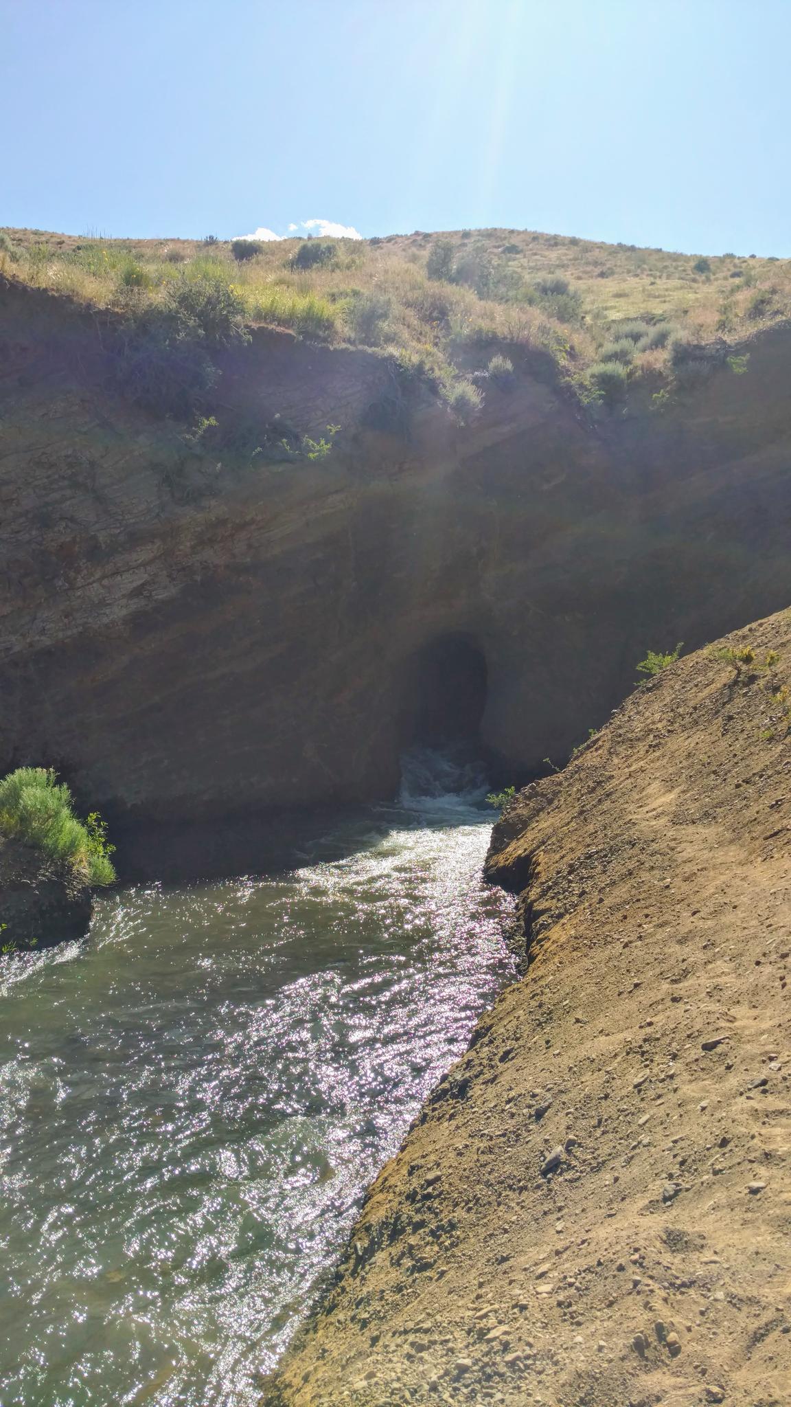 River flowing into a dark cave opening under a sunny, dry hillside.