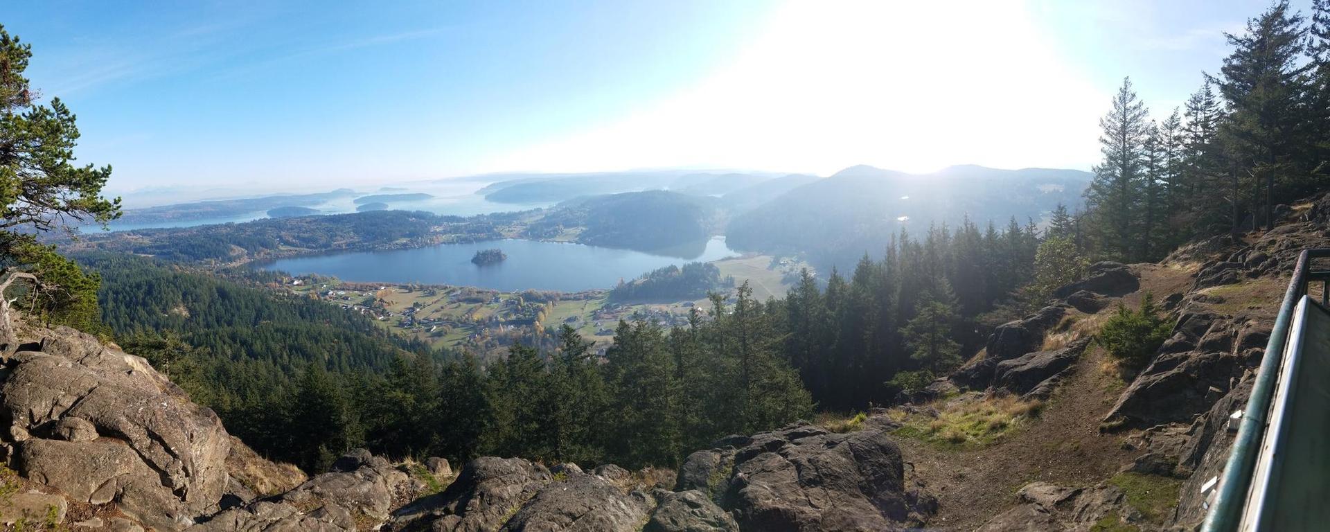 Panoramic view of a large lake surrounded by forests and hills on a sunny day.