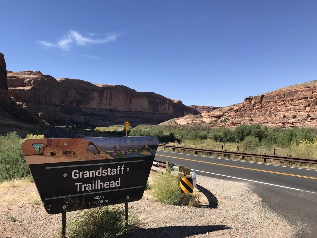 Trailhead sign by a road, with distant red rock cliffs under a clear sky.