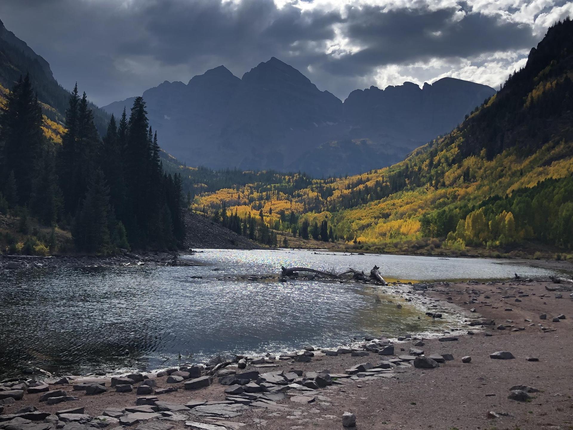 Dark lake reflects mountains and golden autumn trees under a cloudy, dramatic sky.